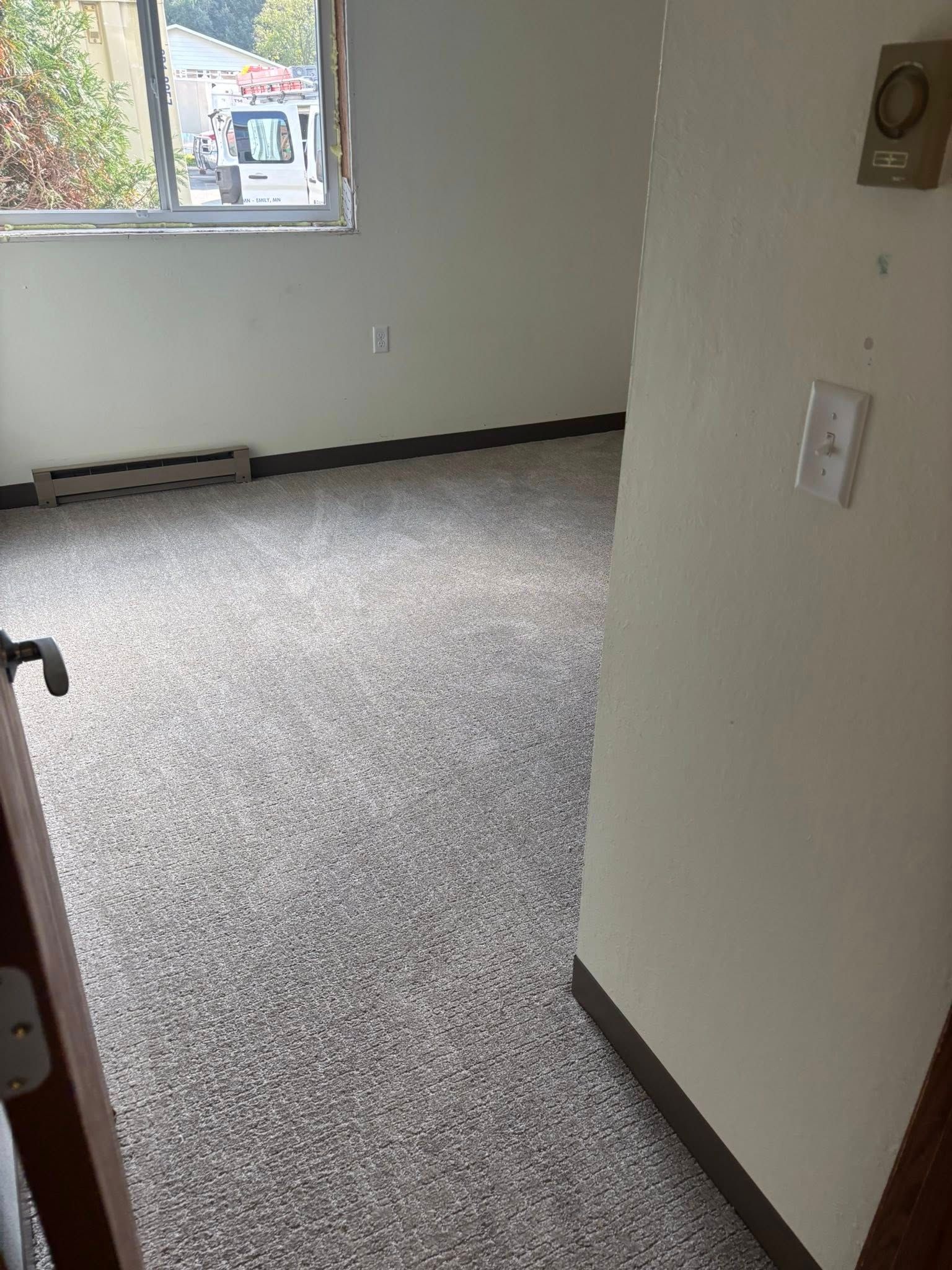 An interior view of an empty bedroom with light-colored walls, speckled gray carpet, and a window overlooking a street.