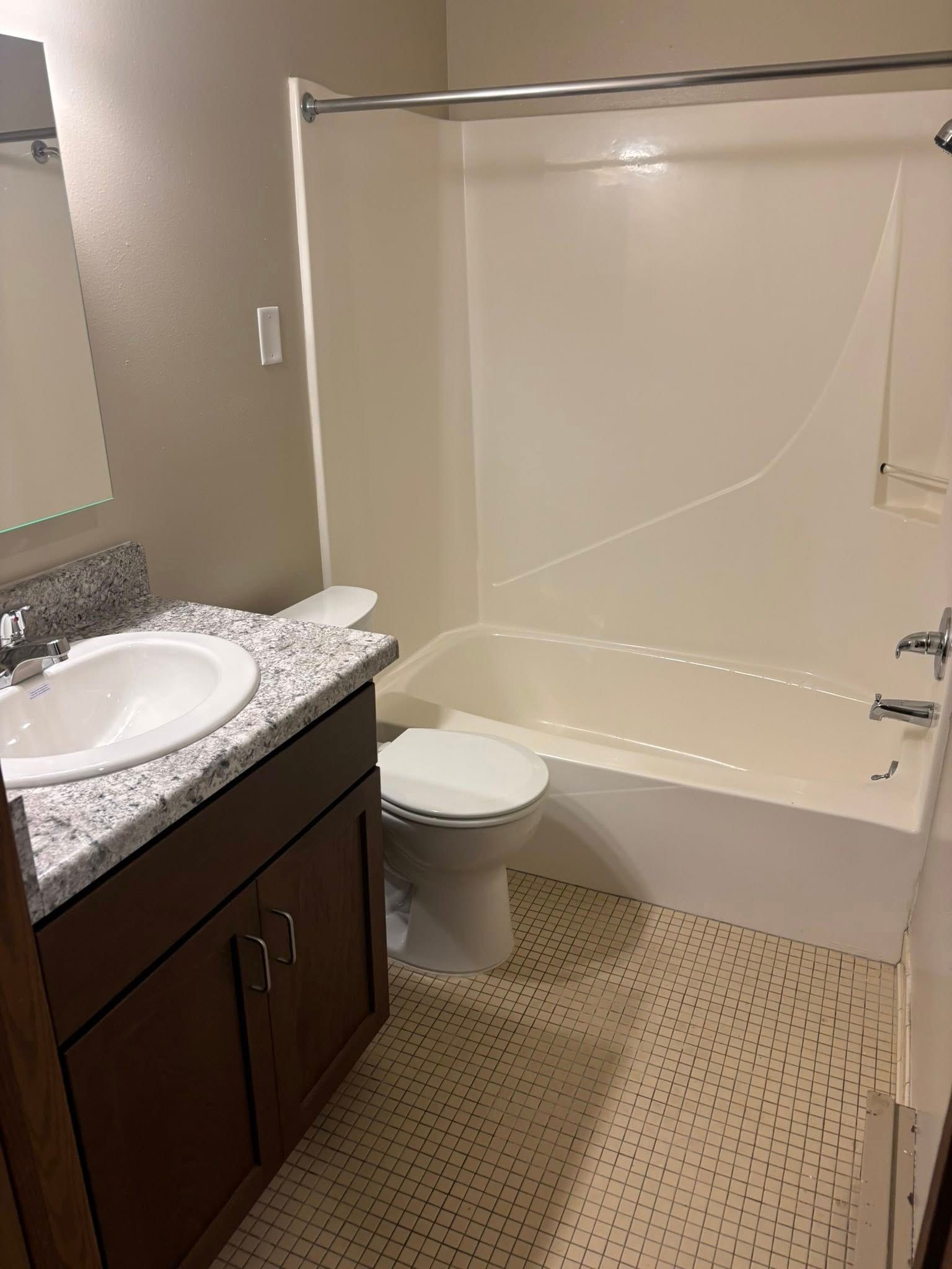 A bathroom with a white tub and shower, a toilet, and a vanity with a granite-style countertop and dark wood cabinets.