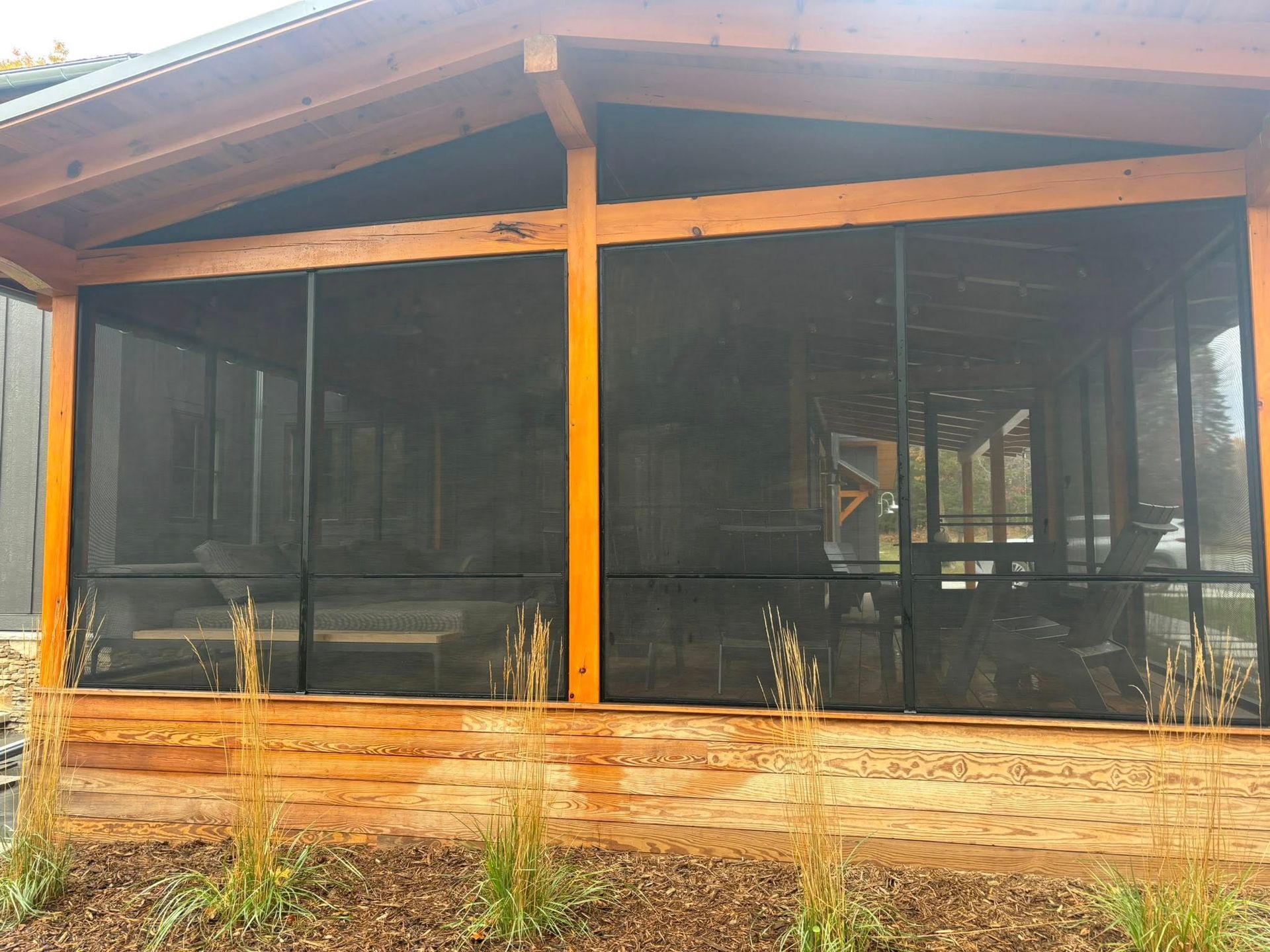 A wooden-framed screened-in porch with interior seating, viewed from the exterior with small grasses in the foreground.