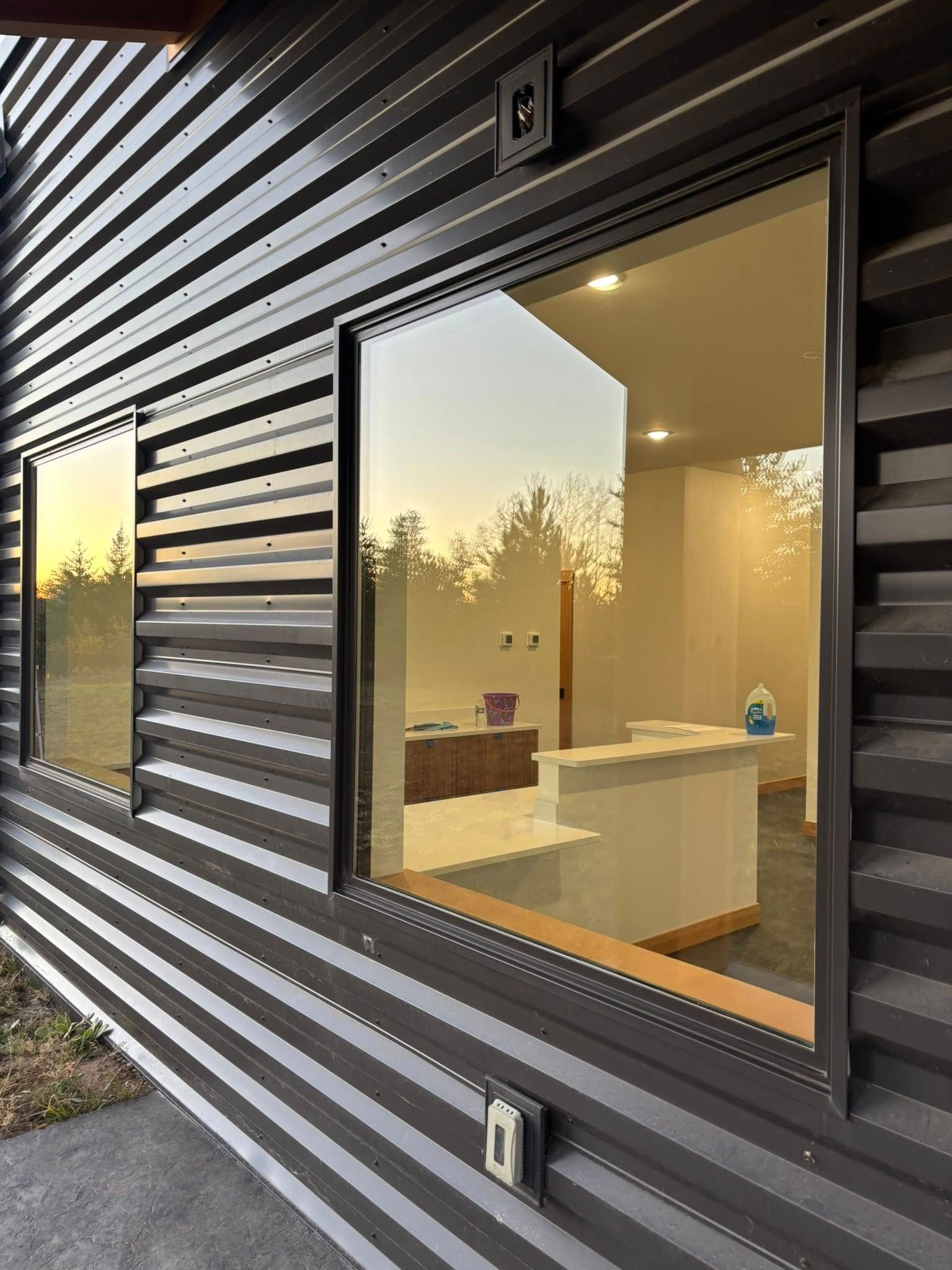 A modern dark corrugated metal building exterior with two large windows revealing a bright, minimalist office interior.