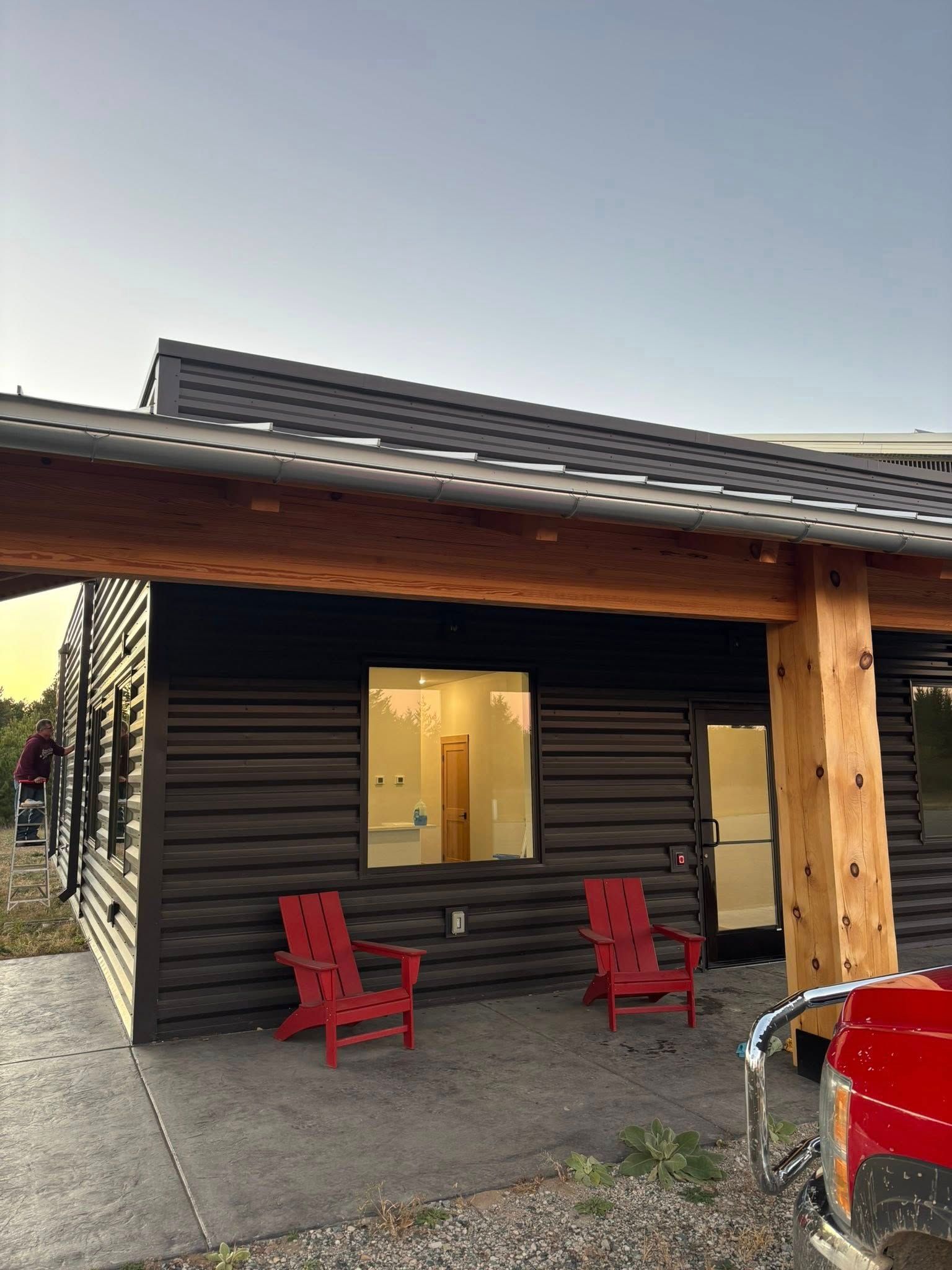A dark horizontal-paneled building with a natural wood porch, large window, and two red chairs on a concrete patio.