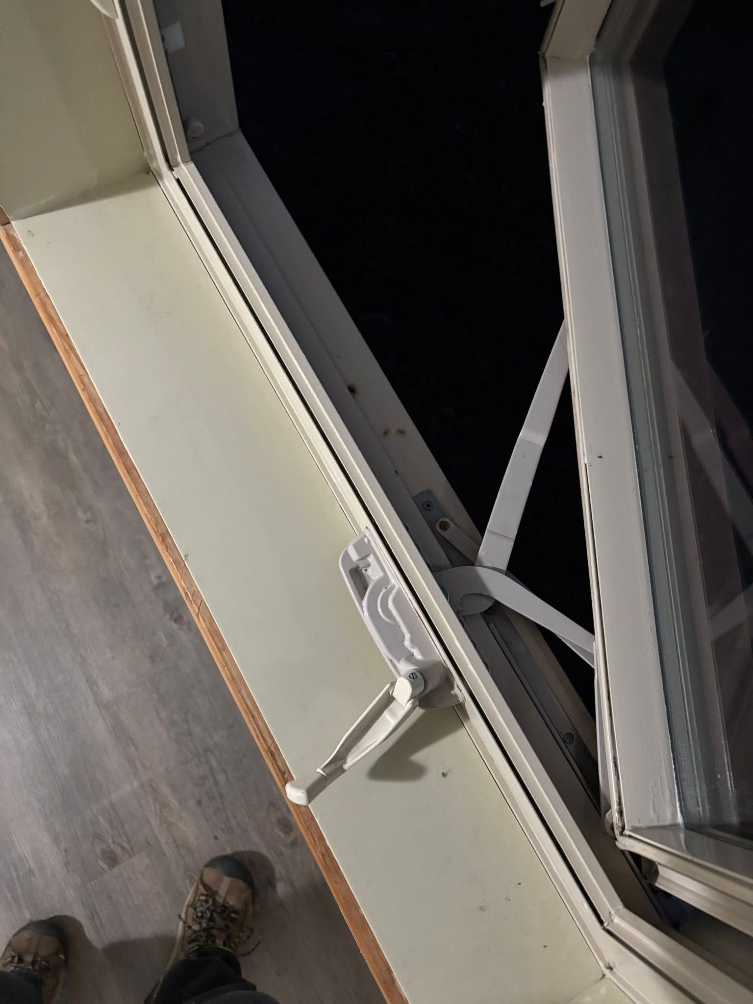 A white casement window is open, showing a dark outdoor night view and a pair of feet wearing boots on a wooden floor.