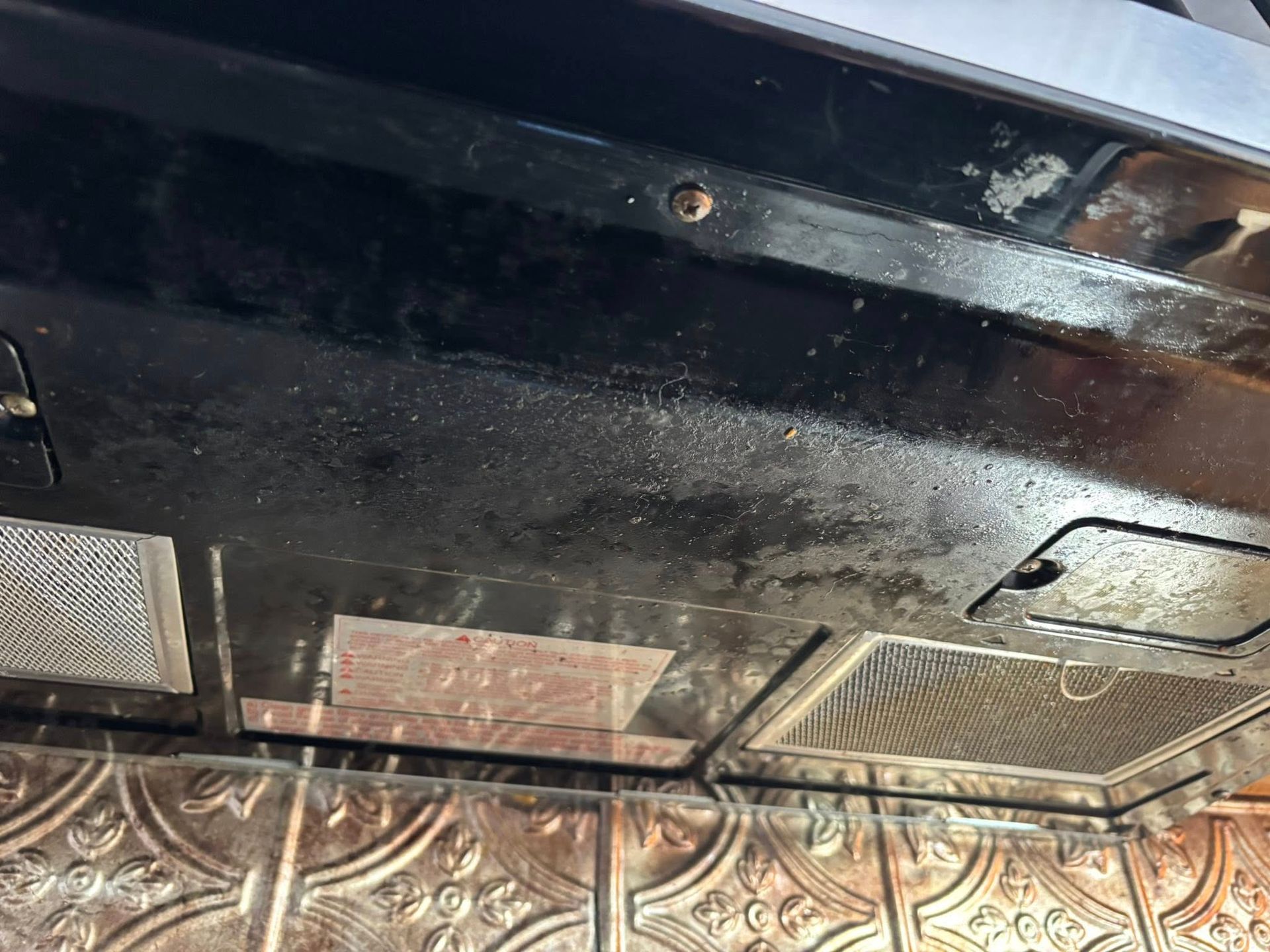 A close-up view of a greasy, black kitchen range hood filter and exterior panel mounted above a decorative tile backsplash.