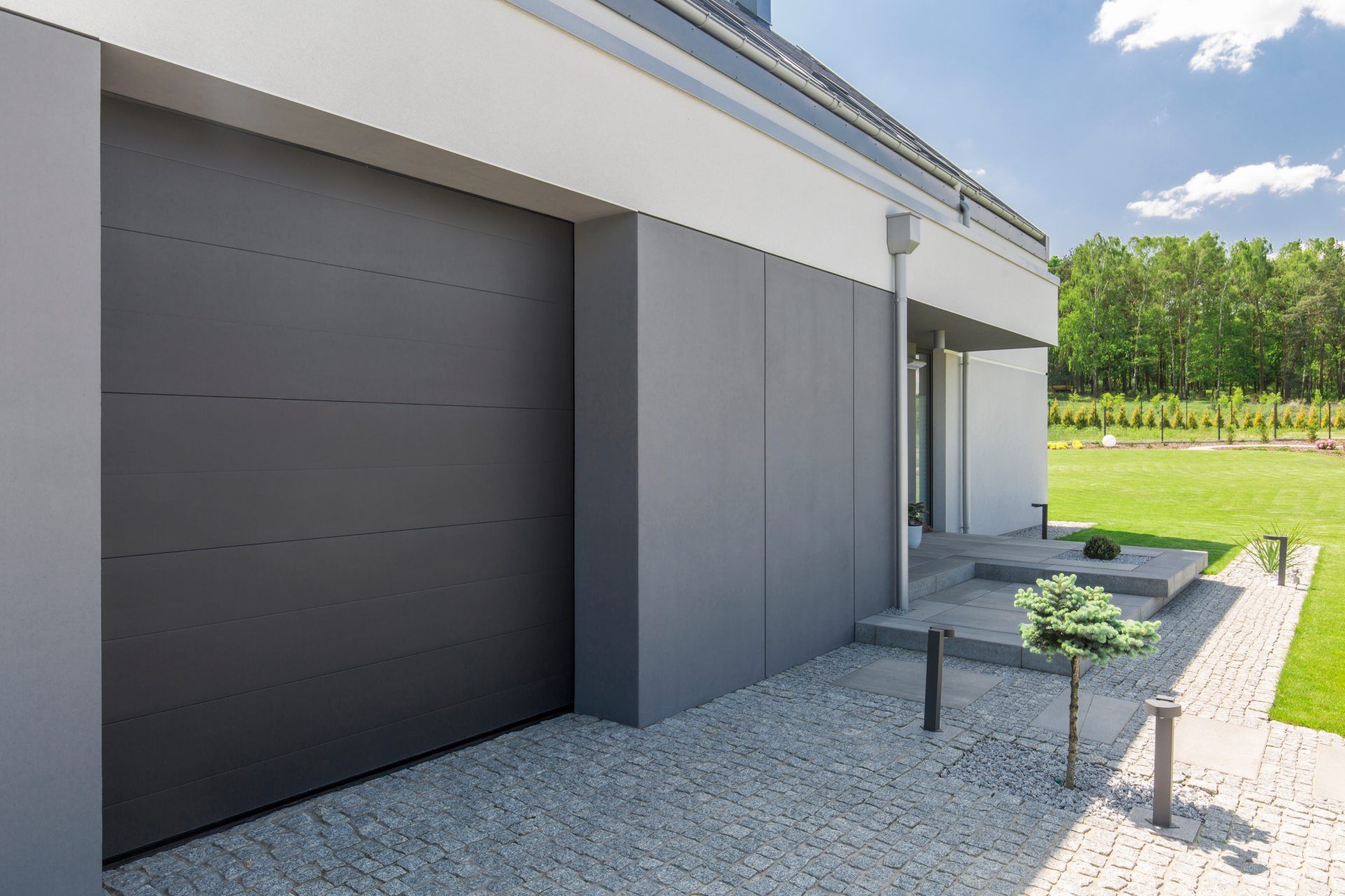 Modern dark gray flush-panel garage door beside a paved patio on a contemporary home.
