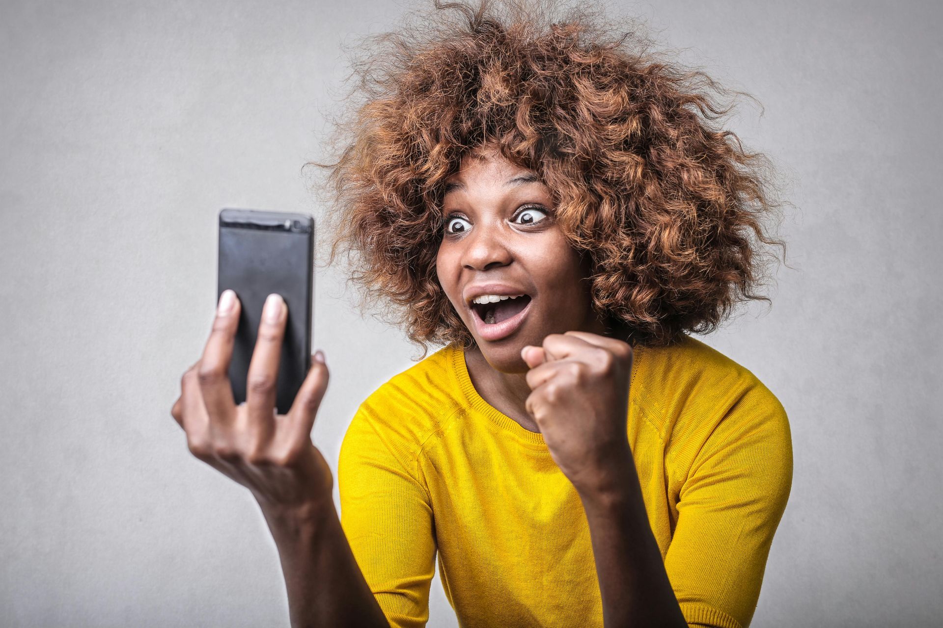 Woman with curly hair, in yellow shirt, looks excitedly at phone held in her hand.