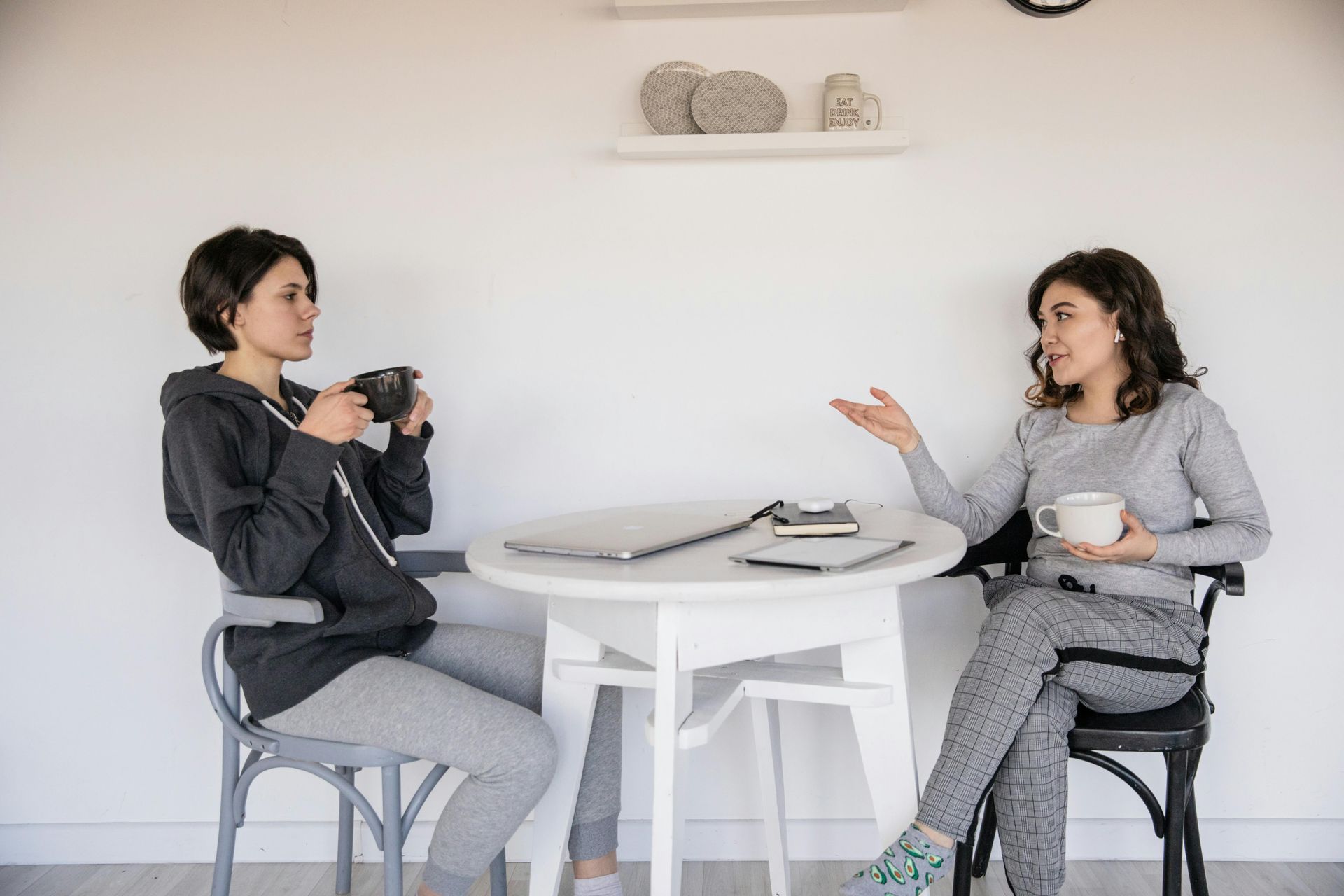 Two people chatting at a small white table, holding mugs in a bright room