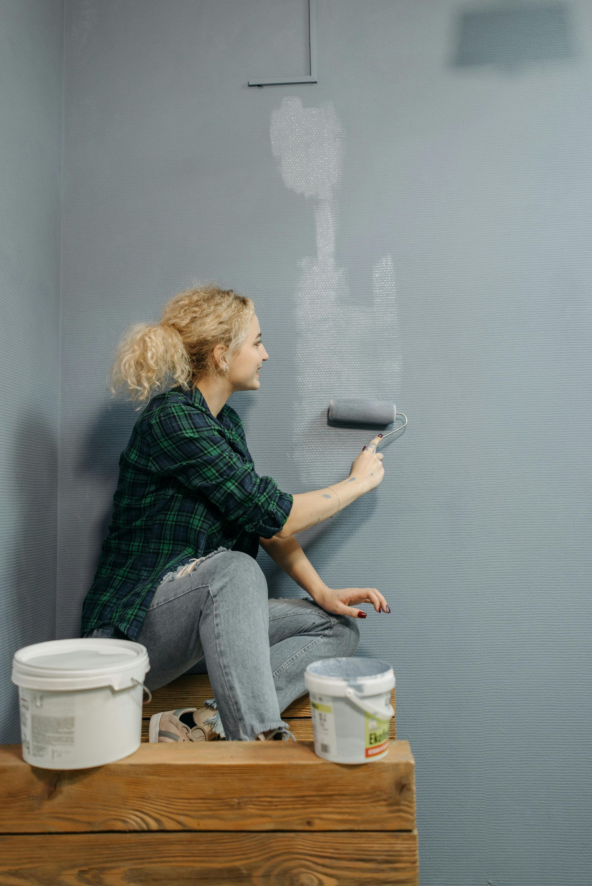 Woman painting a gray wall with a roller, sitting on a wooden ledge, smiling.