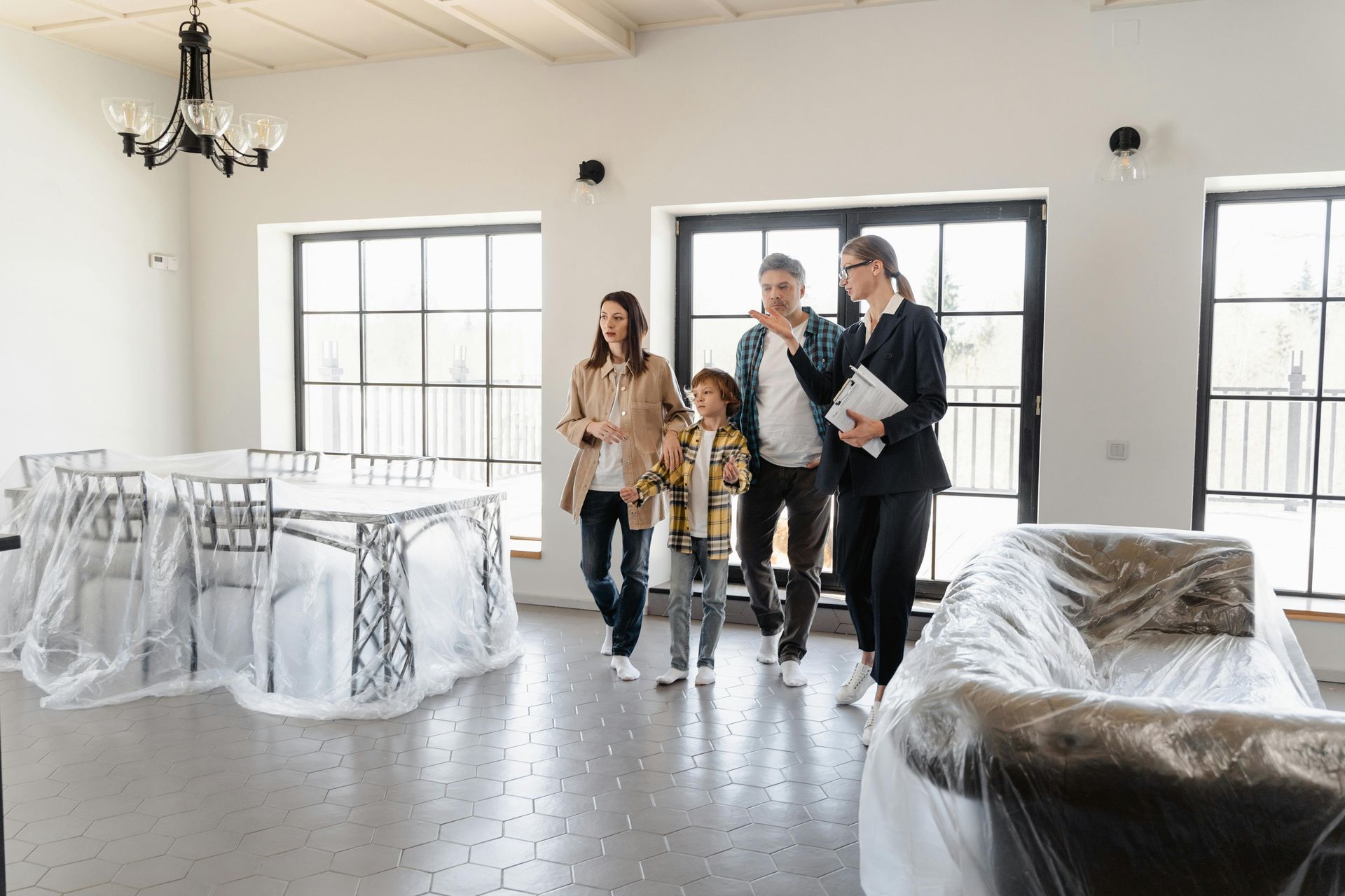 Four people touring a bright, unfinished room with white walls, windows, and covered furniture.