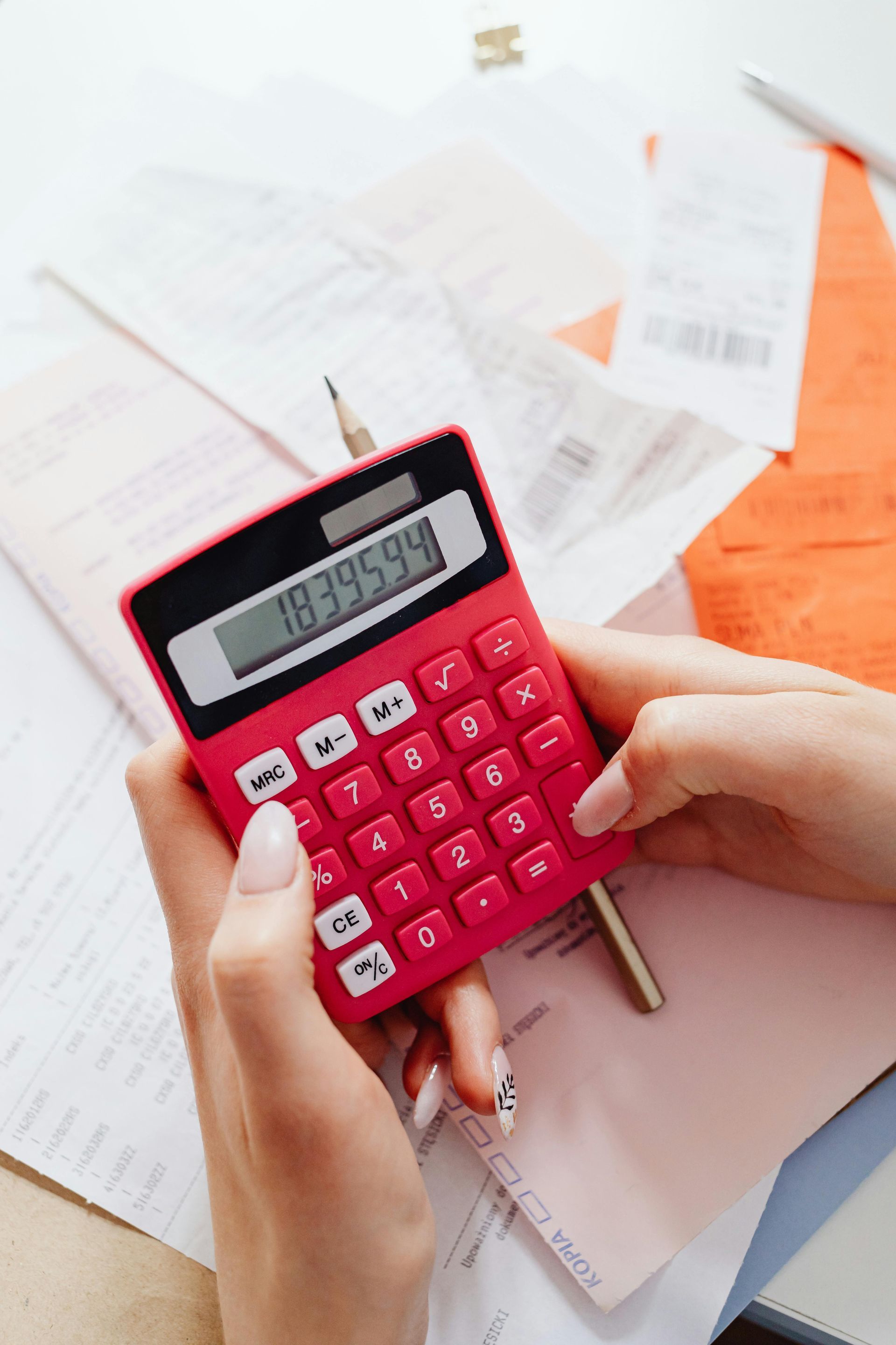 Hands using a red calculator on a desk, surrounded by receipts and papers, calculating finances.
