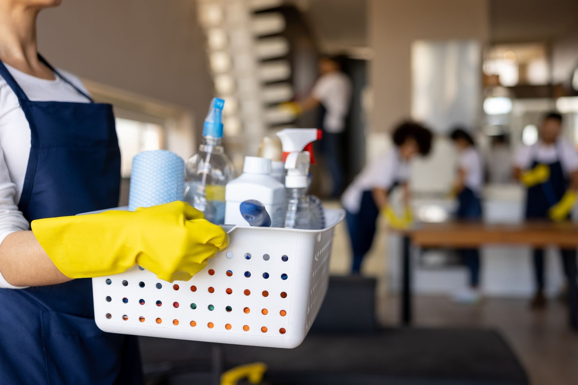 A woman wearing yellow gloves is holding a basket of cleaning supplies.