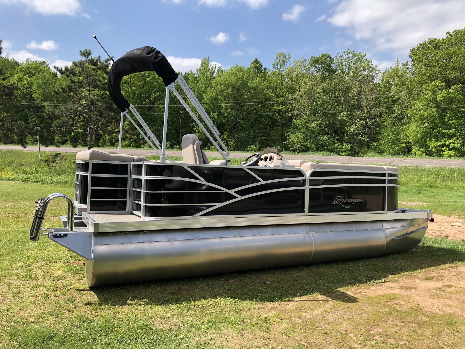 Pontoon boat on grass, under blue sky. Silver hull, black accents, and canopy.