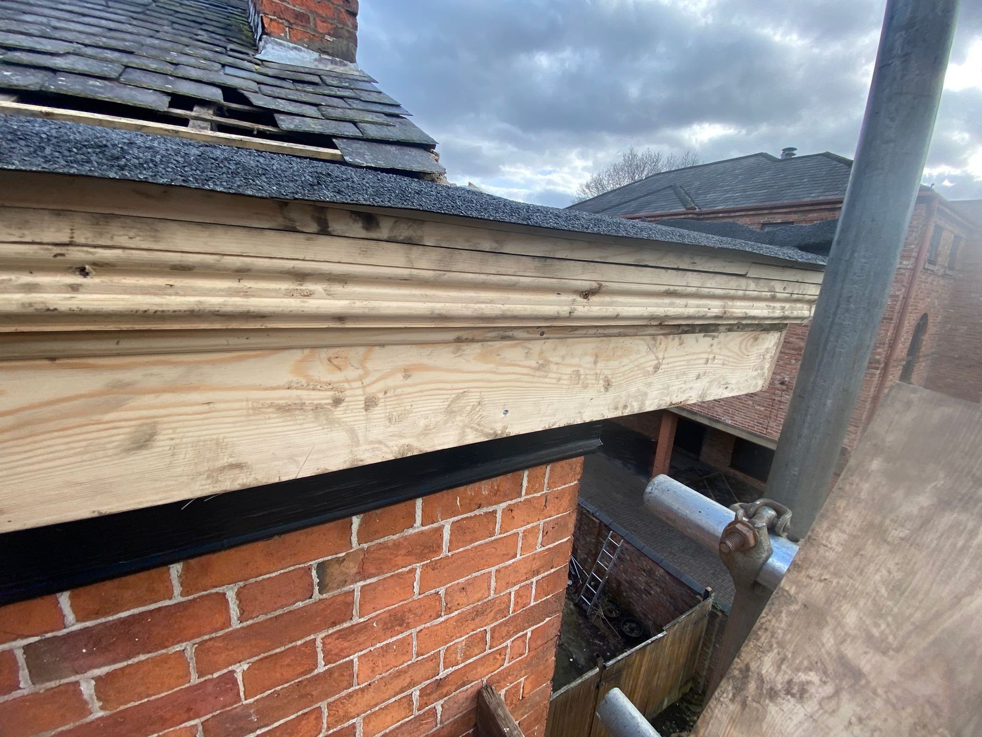 Wooden roof trim being installed on a brick building, with a partially visible slate roof and scaffolding.