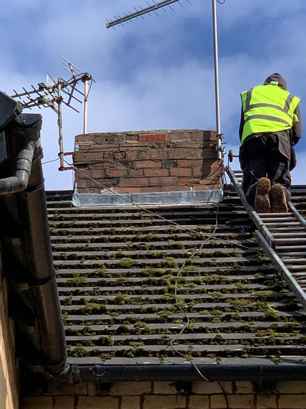 Person in high-visibility vest on ladder, working on a brick chimney with an antenna, on a rooftop.