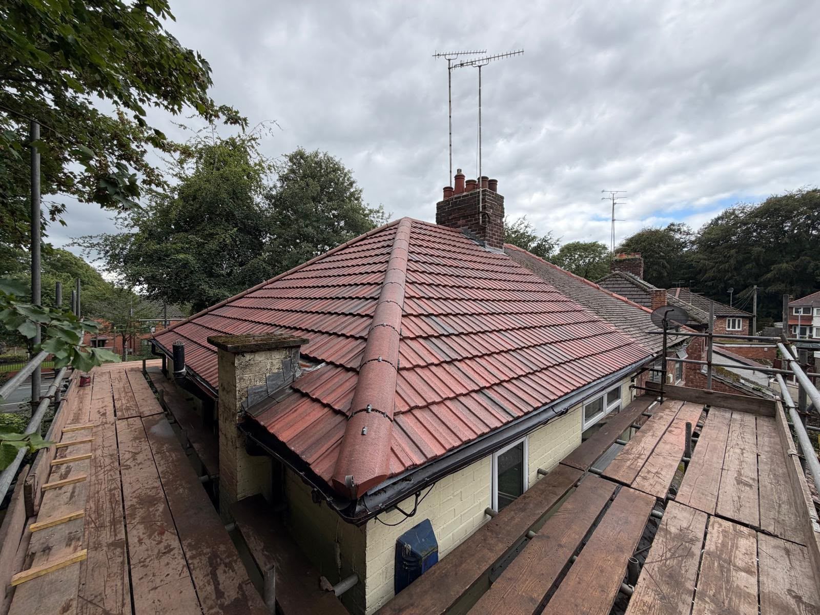 Rooftop view of a house with red tiles, chimneys, and scaffolding under a cloudy sky
