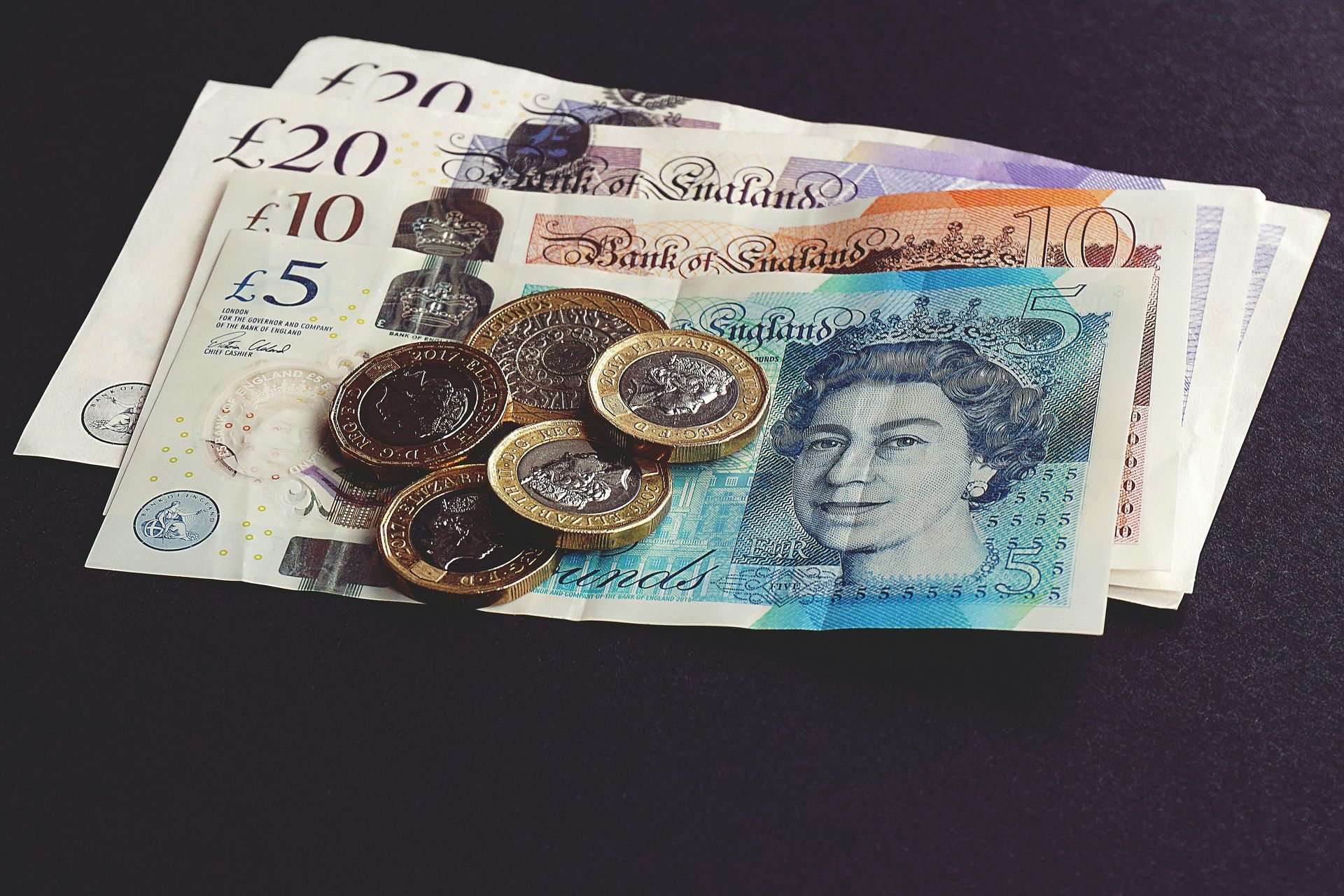 Pile of British banknotes and coins on a dark surface. Currency includes £5, £10 and £20 notes, and various coins.