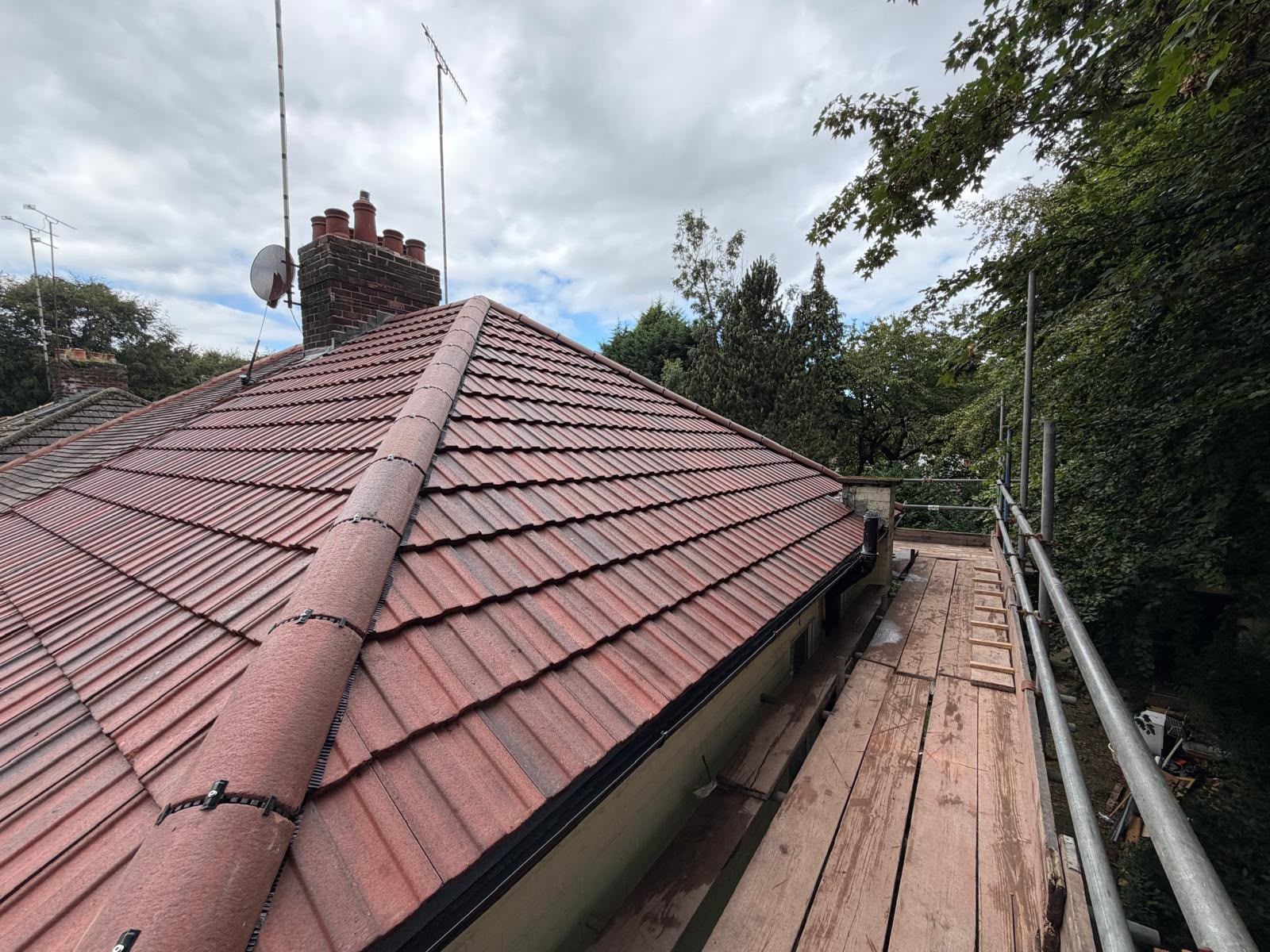 Red tiled roof with chimney beside a narrow wooden walkway and trees under a cloudy sky