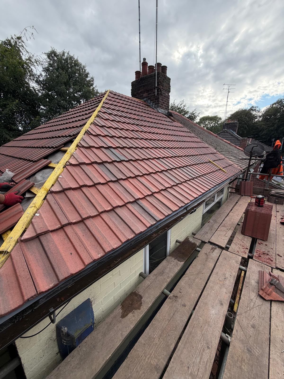 Roof tilers working on a red-tiled sloped roof beside scaffolding and a brick chimney under cloudy skies