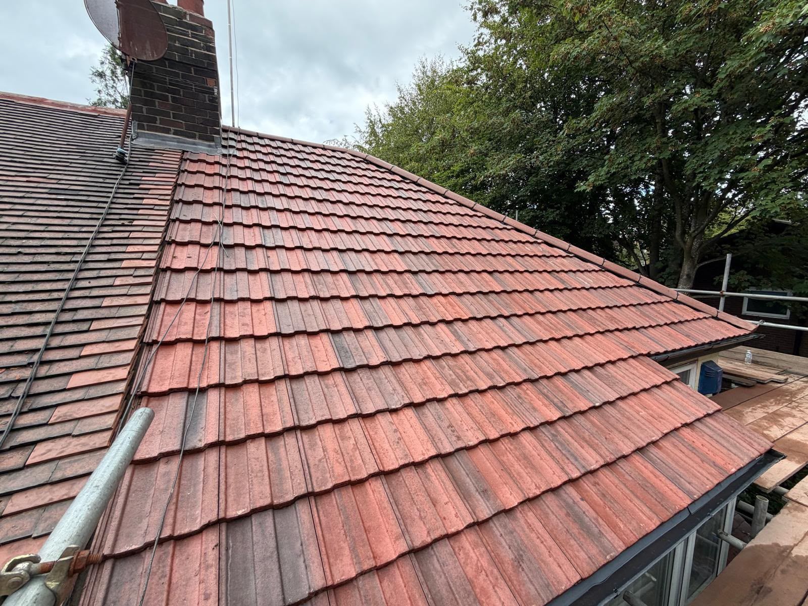 Red-tiled sloped roof with a chimney, viewed from above against trees and cloudy sky
