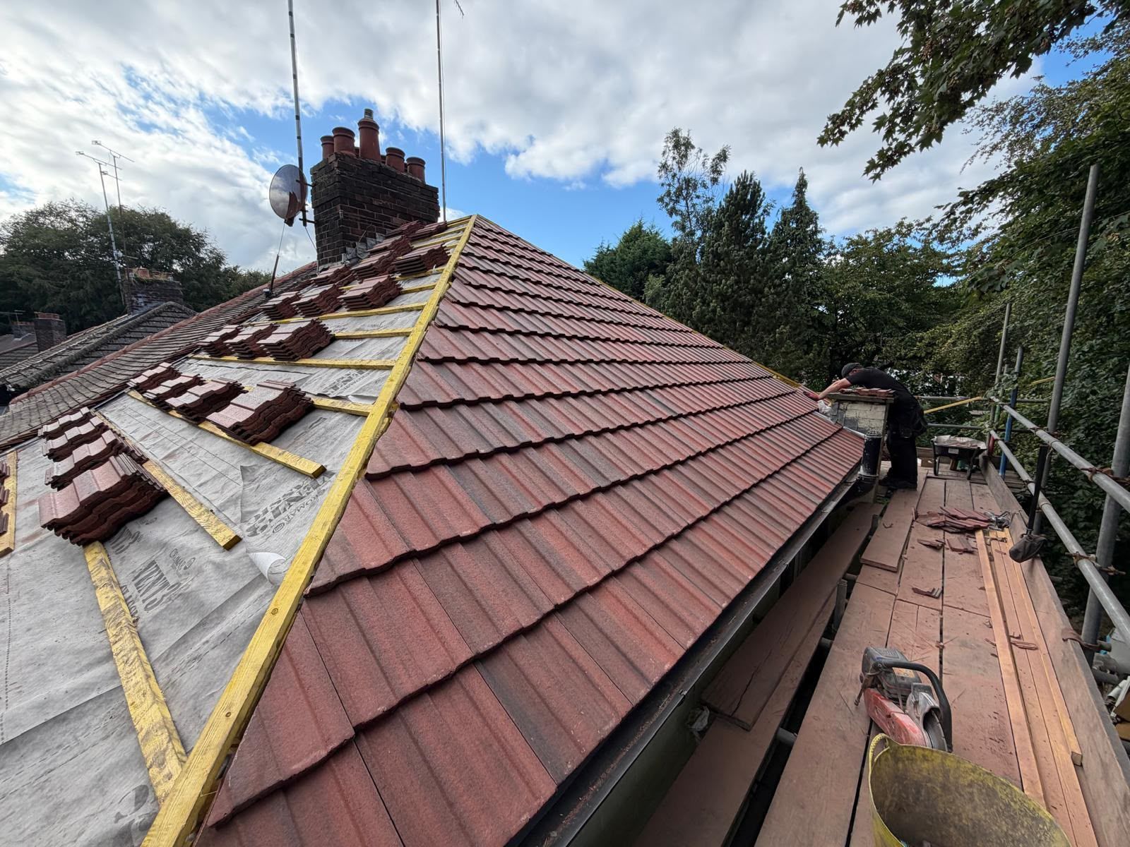 Roofers working on a red-tiled house roof with yellow safety harness lines under a cloudy sky