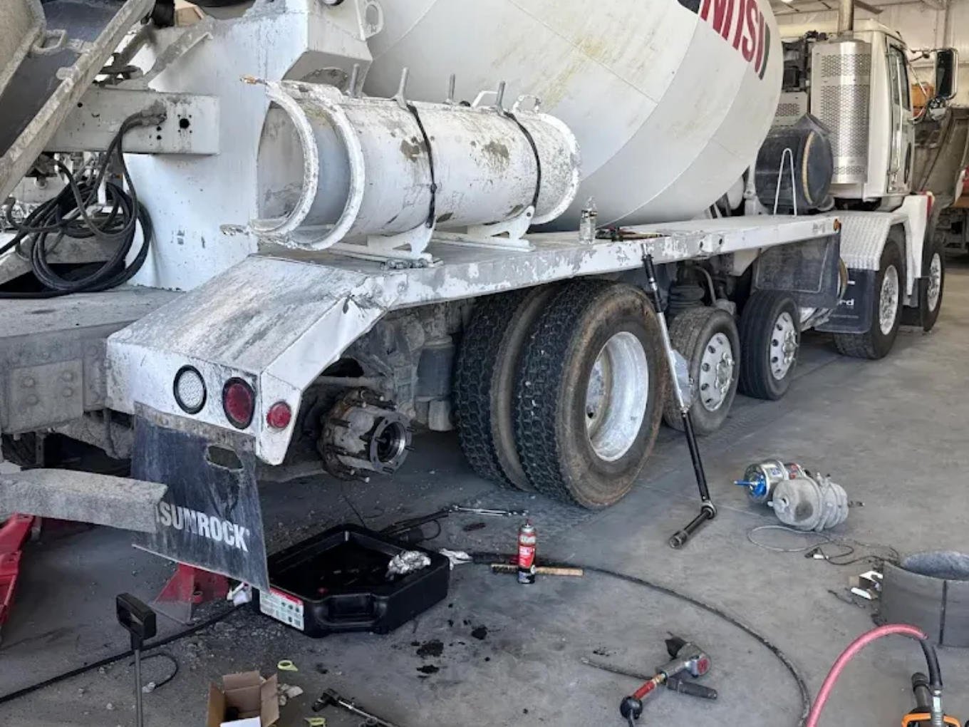 Cement truck undergoing maintenance, wheels removed, tools and oil pan on the ground, likely in a garage.