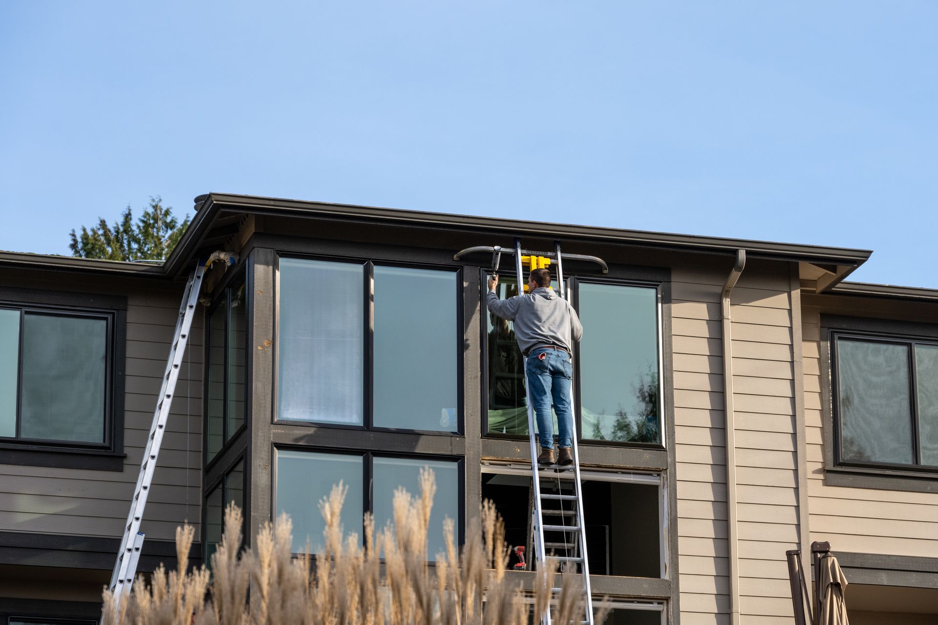 Man on ladder washing windows of a two-story beige house under a blue sky.