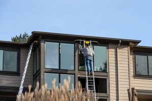 Une personne sur une échelle nettoie de grandes fenêtres d'une maison au bardage beige et au toit marron.
