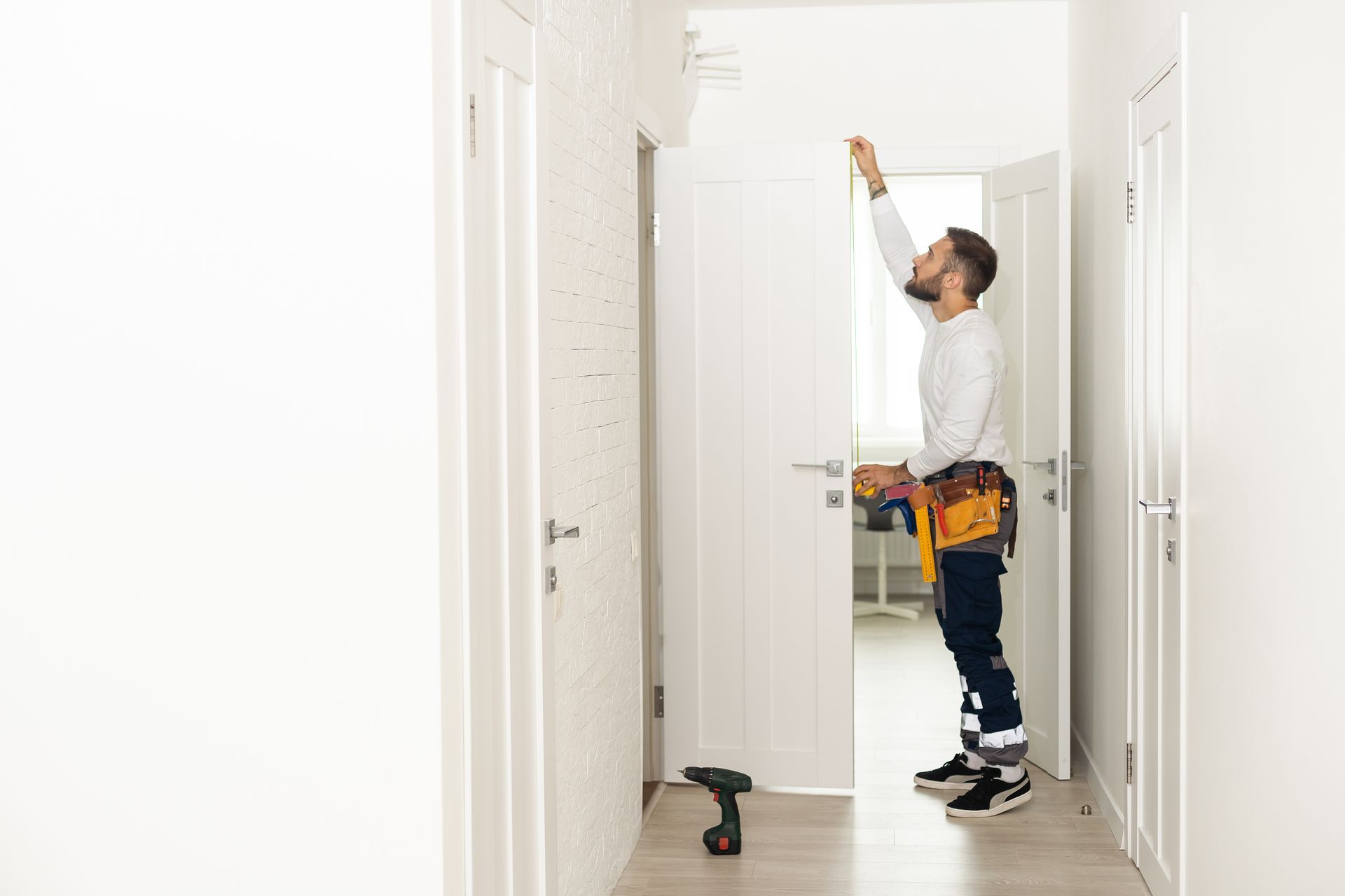 Man with tool belt installs something above a door in a white hallway.