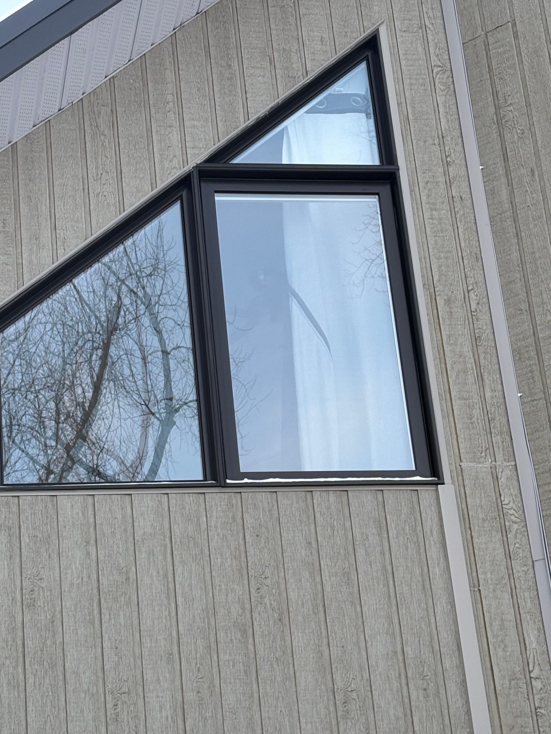 Fenêtre triangulaire à cadre noir sur un immeuble en béton gris. Reflets des arbres et du ciel visibles.