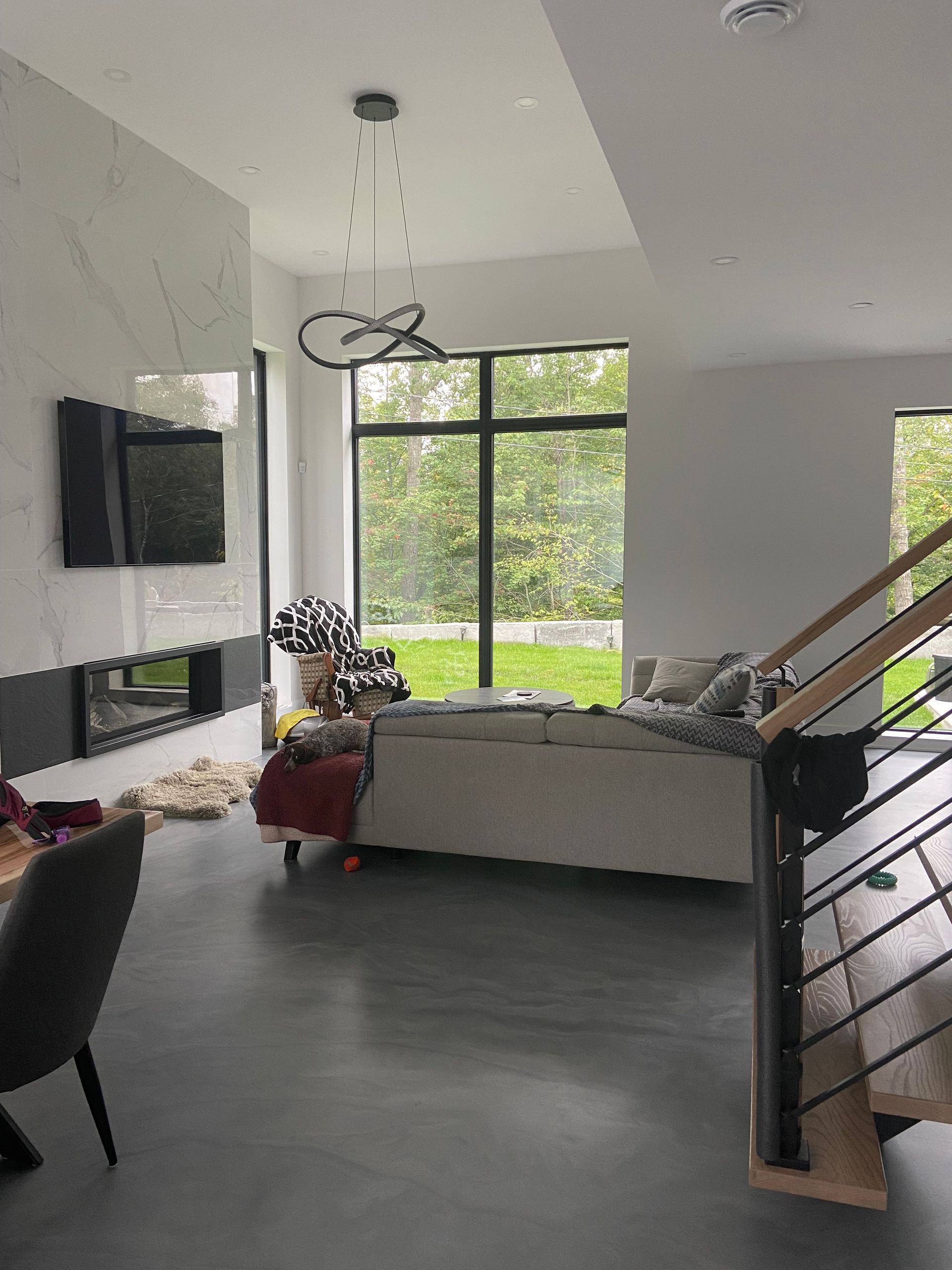 Salon moderne avec cheminée, grande fenêtre et escalier, sol gris, canapé beige et luminaire suspendu.