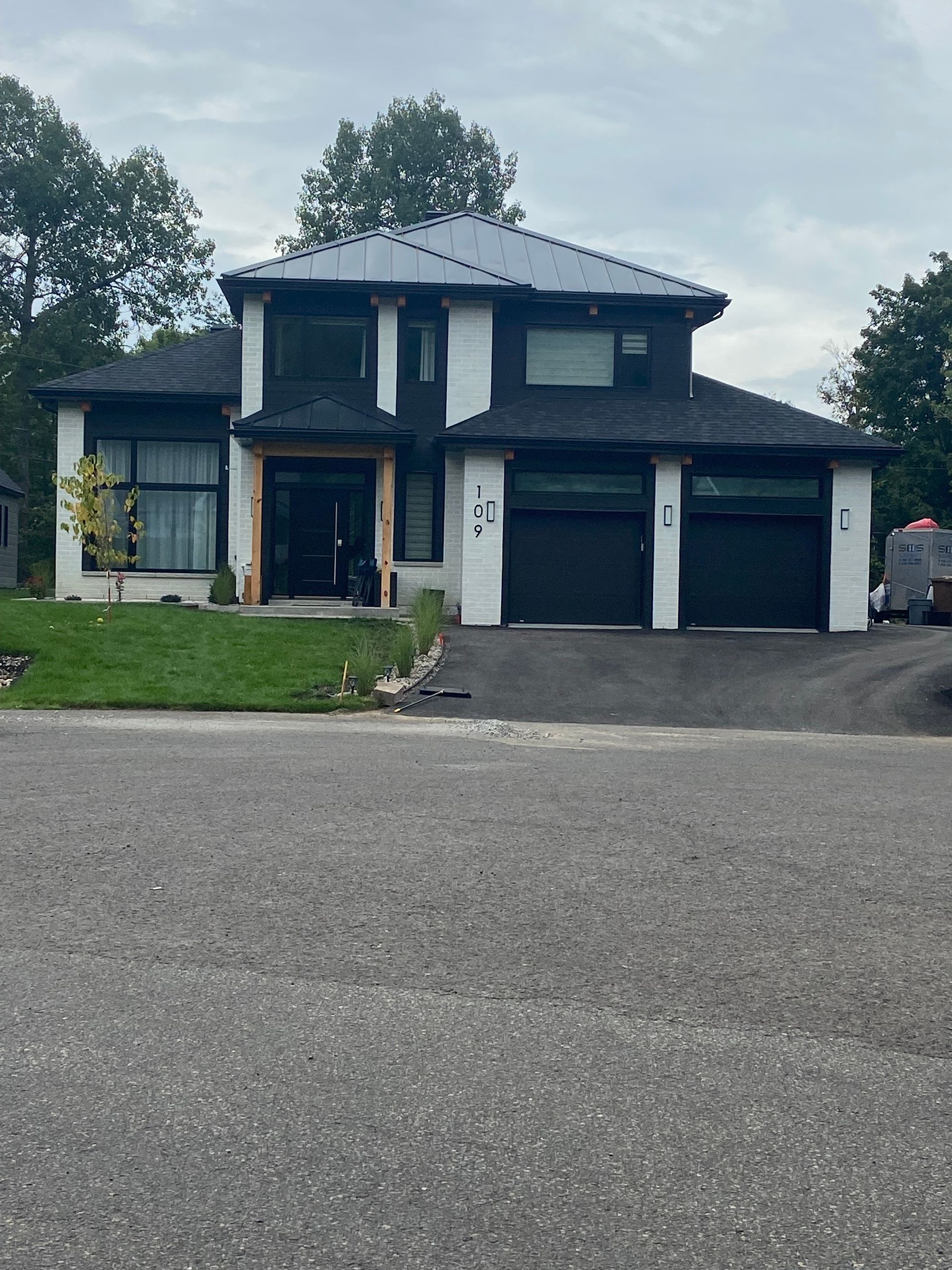 Maison moderne à deux étages avec boiseries noires, bardage blanc et garage pour trois voitures. Ciel couvert.