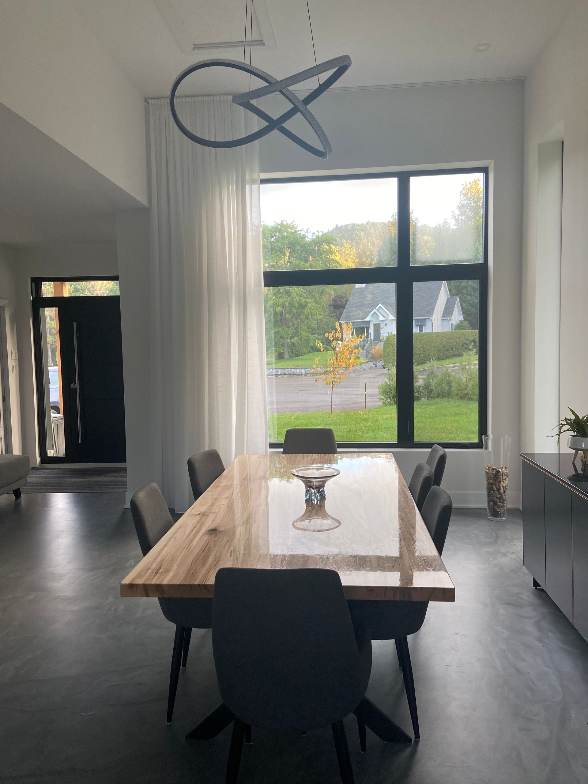 Salle à manger avec longue table en bois, chaises grises, grande fenêtre donnant sur le jardin et luminaire moderne.