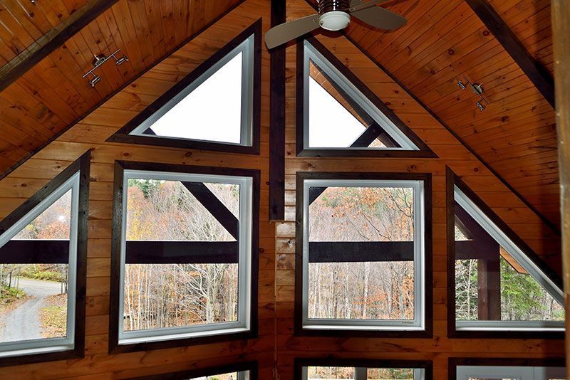 Vue intérieure avec fenêtres triangulaires et rectangulaires, vue extérieure boisée, plafond et murs en bois.