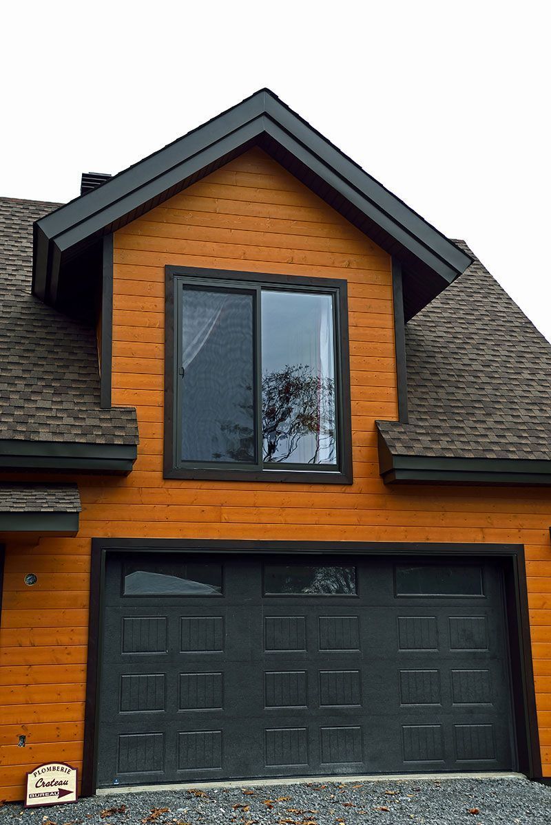 Maison aux boiseries orange, avec porte de garage et fenêtre gris foncé. Des moulures noires soulignent le toit et les fenêtres.