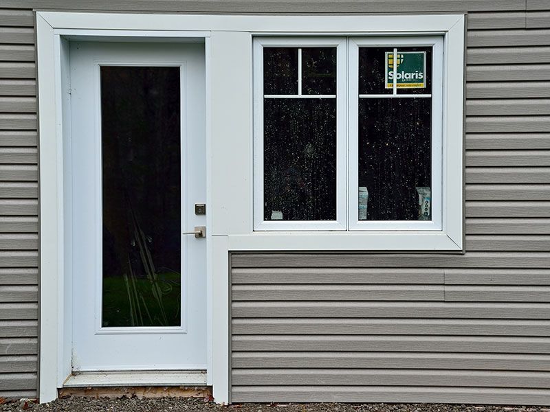 Extérieur d'un bâtiment avec une porte et une fenêtre à cadre blanc. Revêtement extérieur gris.