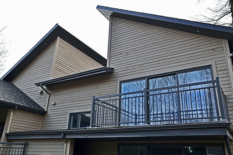 Maison beige avec des boiseries noires, un balcon et de grandes fenêtres reflétant les arbres.