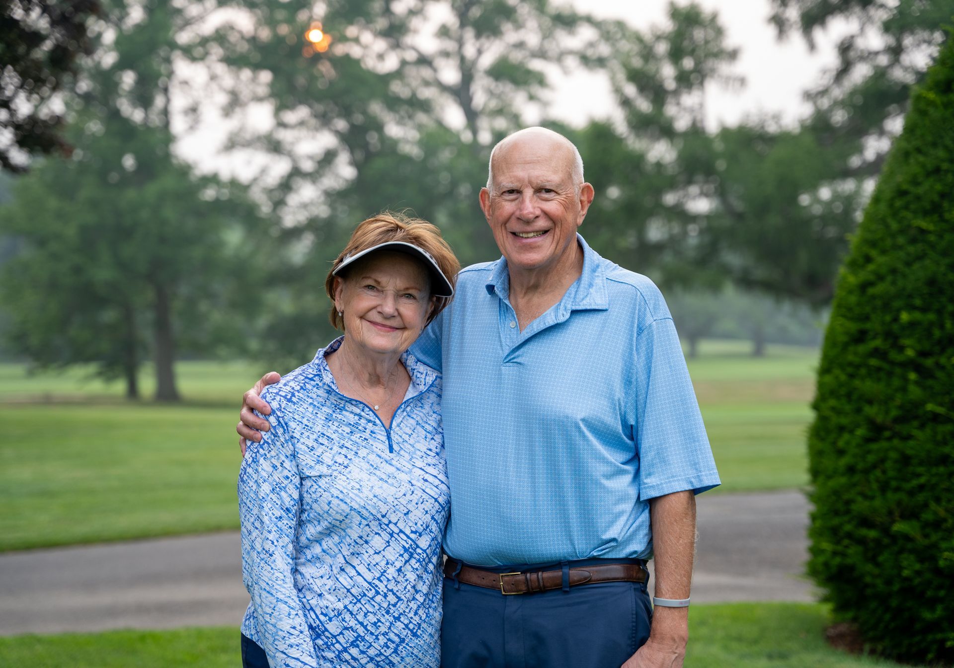 An older couple poses for a photo outdoors, standing side-by-side with arms around each other in a grassy, wooded area.