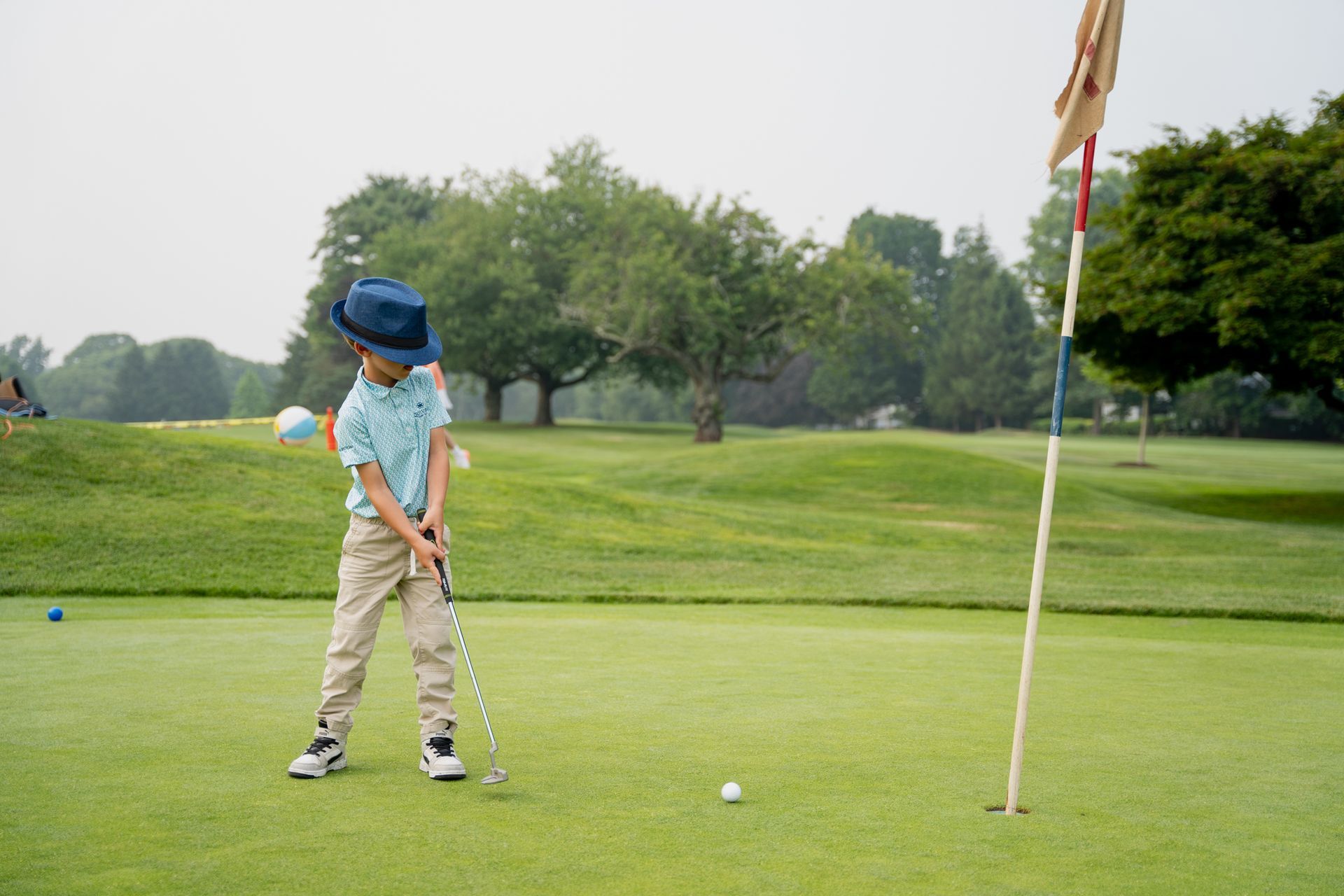 A young golfer in a blue hat and light shirt prepares to putt on a green next to a flagstick.