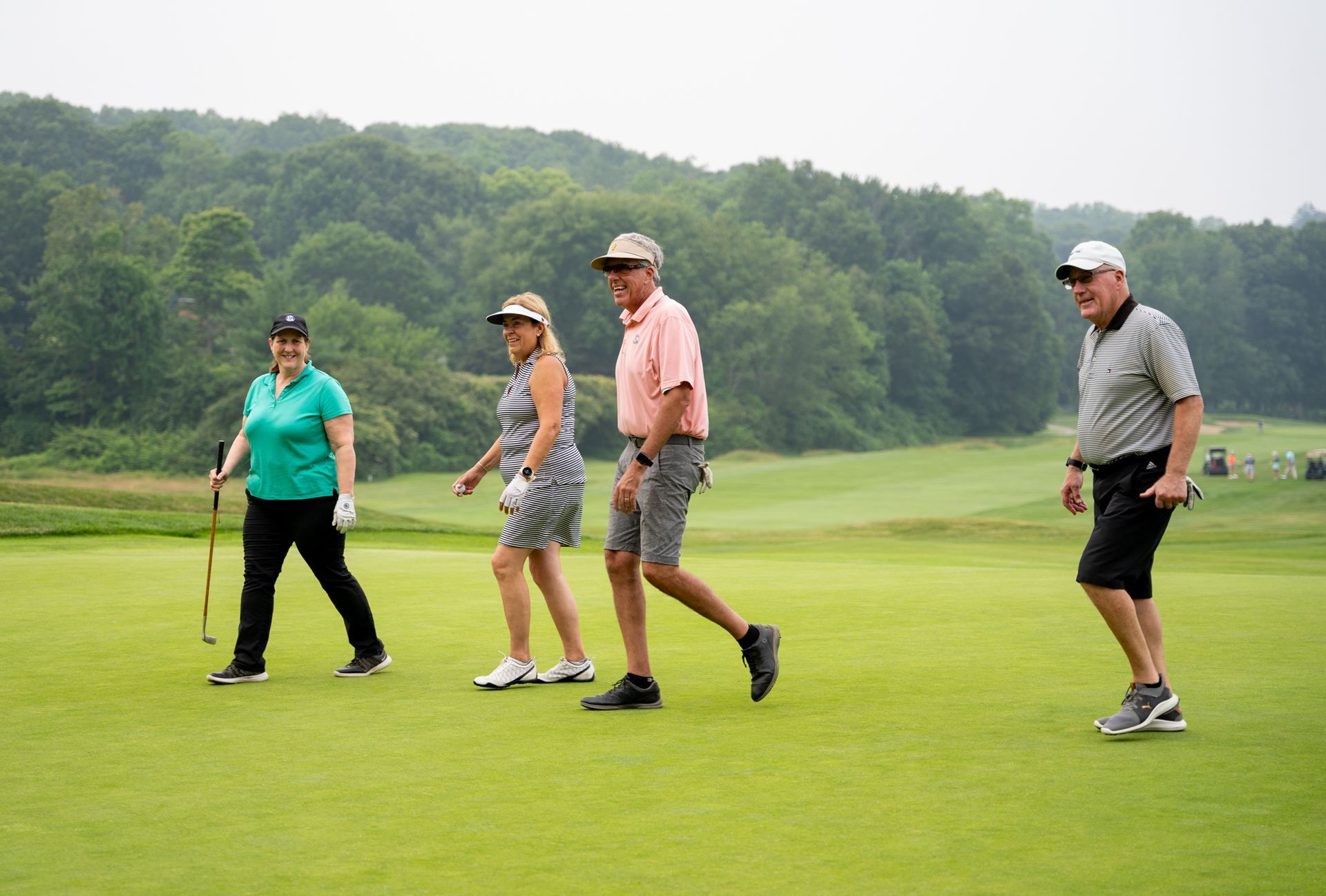 Four people walk across a lush green golf course, smiling and dressed in activewear against a backdrop of trees.