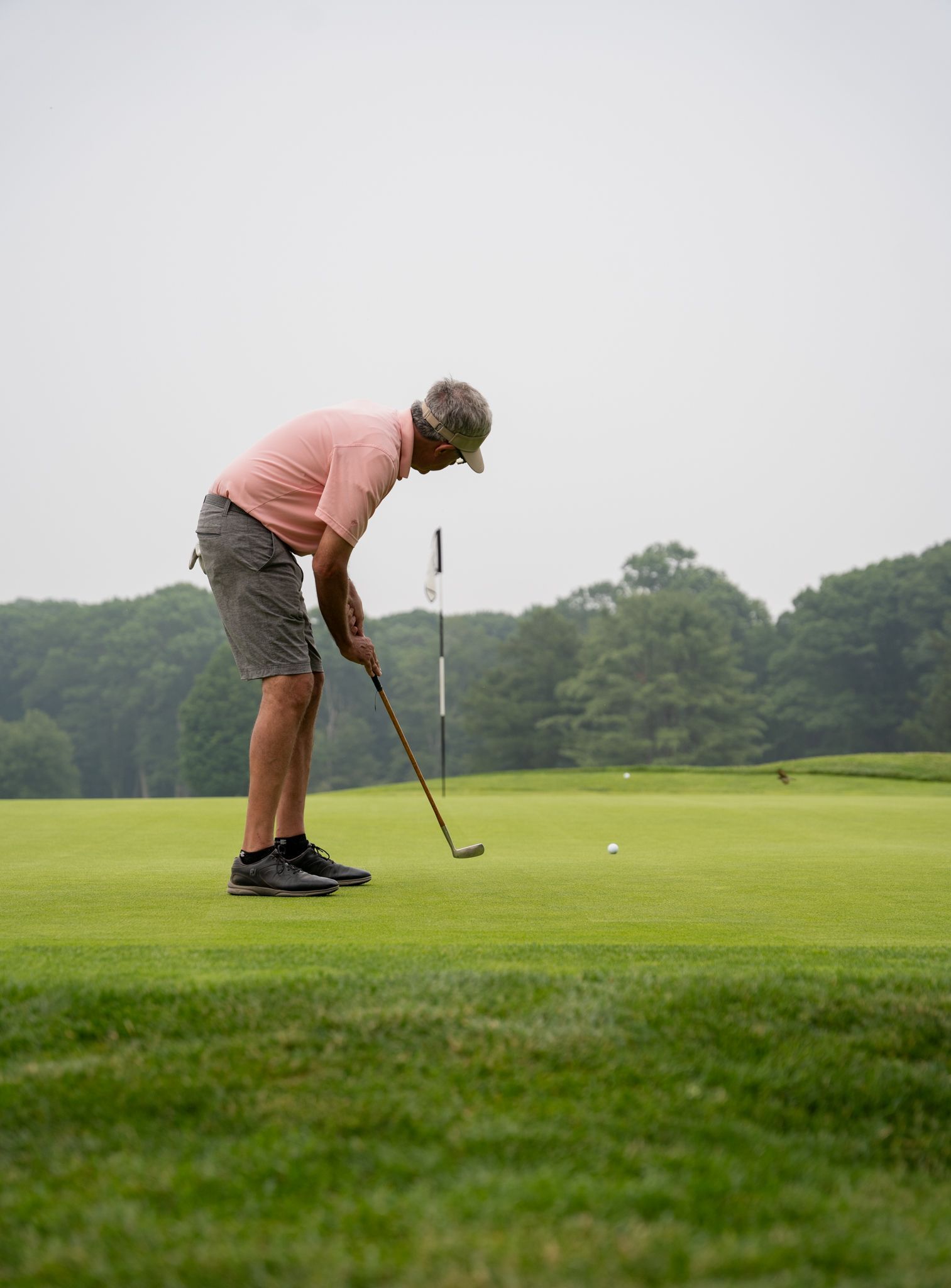 A golfer in a pink shirt and patterned shorts stands on a green, preparing to putt towards a hole in the distance.