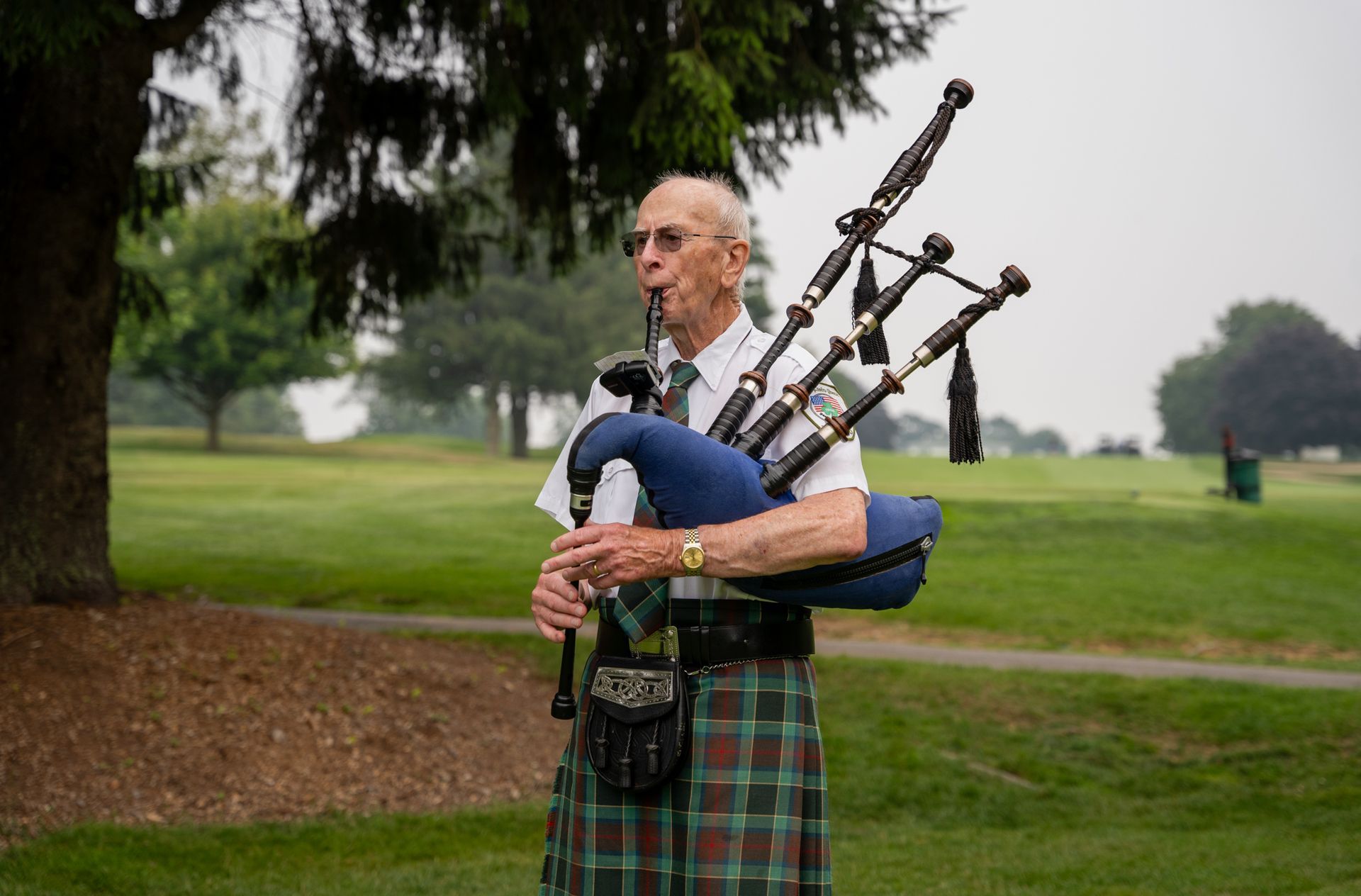 A bagpiper in a kilt plays bagpipes outdoors on a golf course.