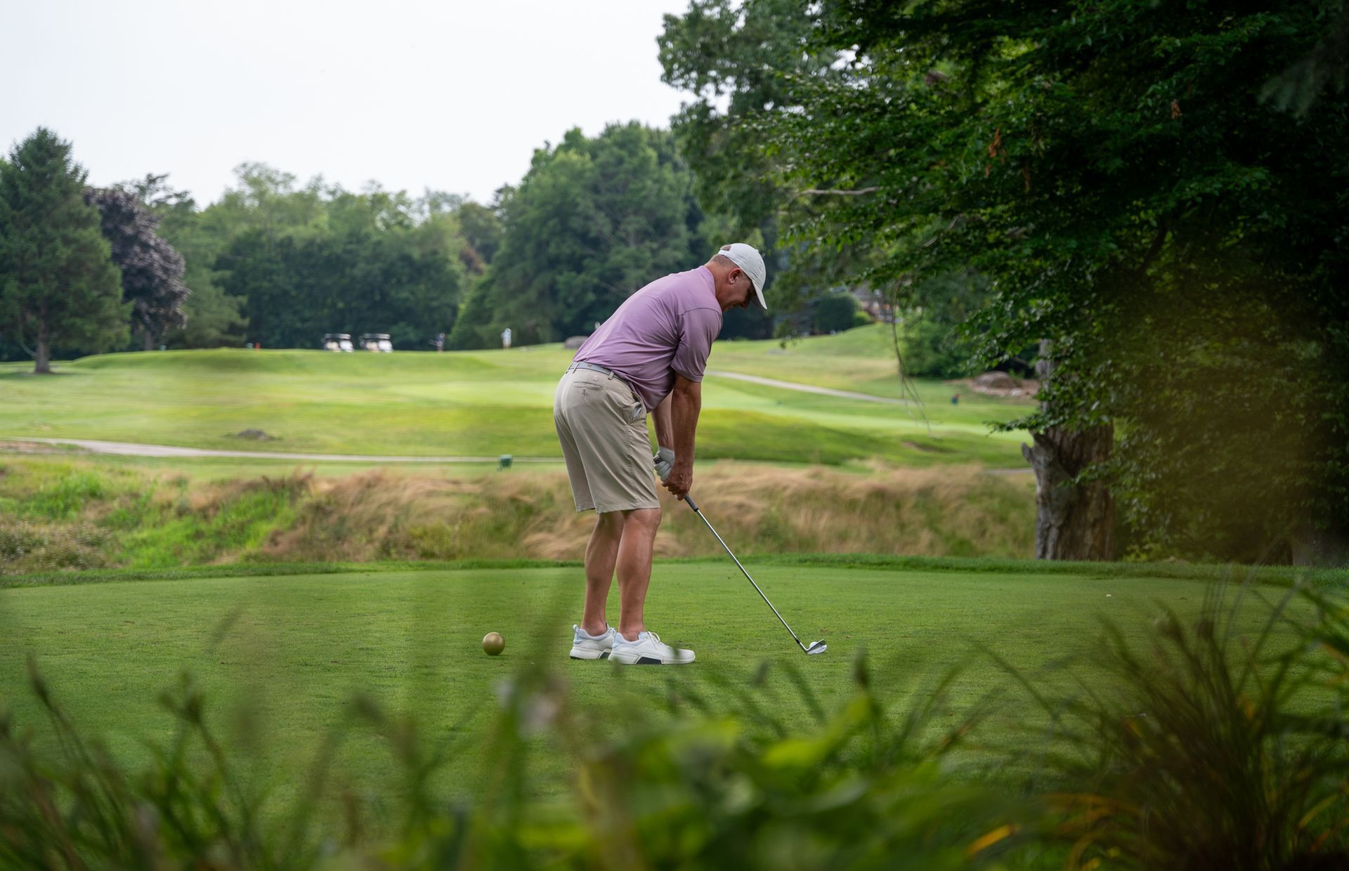 A golfer in a patterned shirt and beige shorts prepares to hit a ball on a lush green course with trees in the background.
