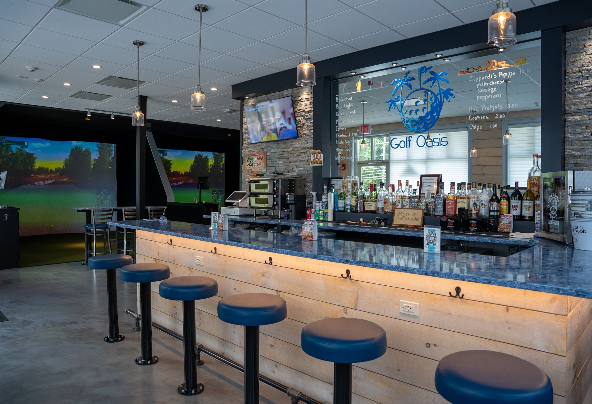A blue-topped bar with five stools in front of a mirrored wall with a logo, next to a golf simulator room.