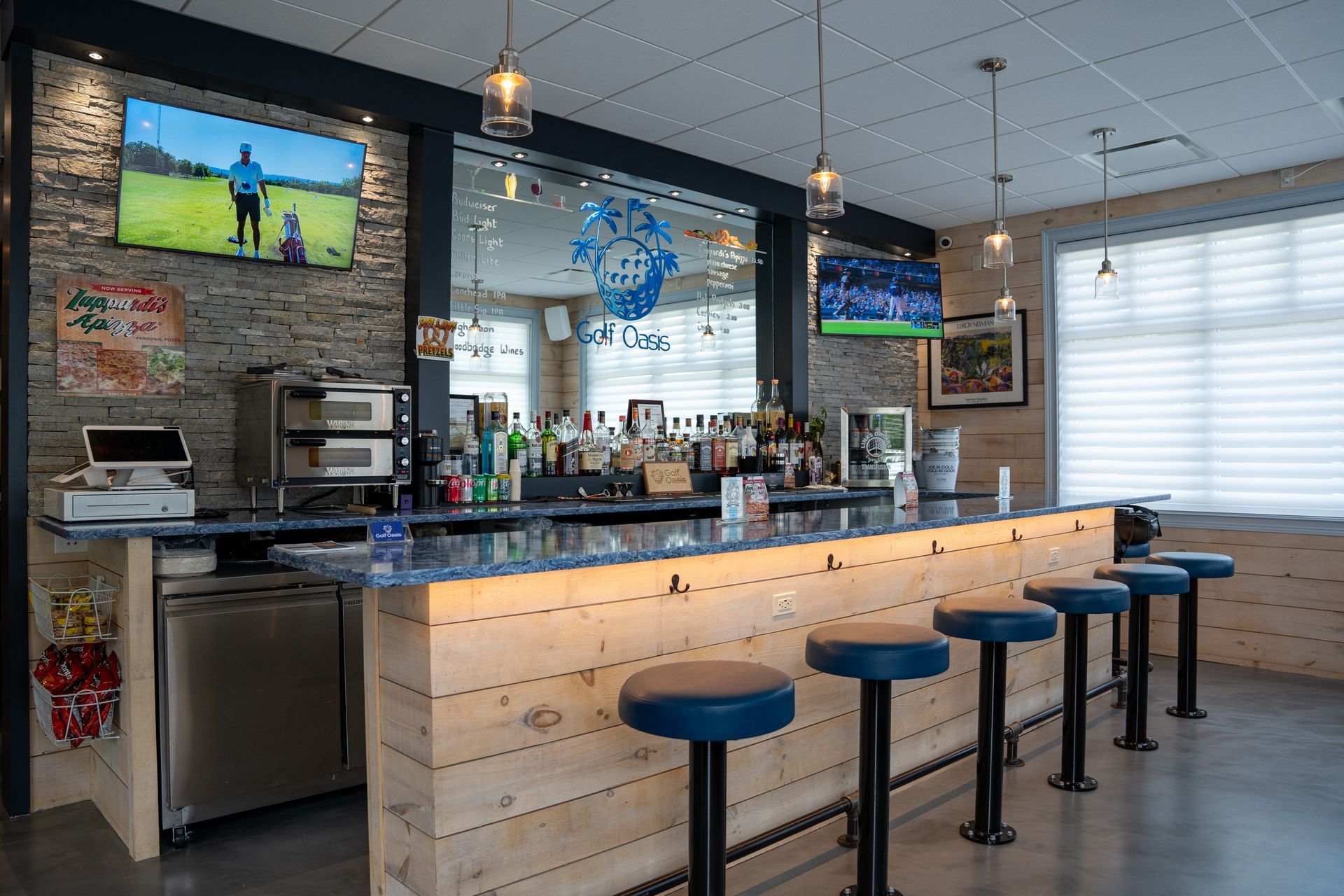 A well-lit bar area with a wooden counter, blue stools, stone accent wall, and TVs showing sports.