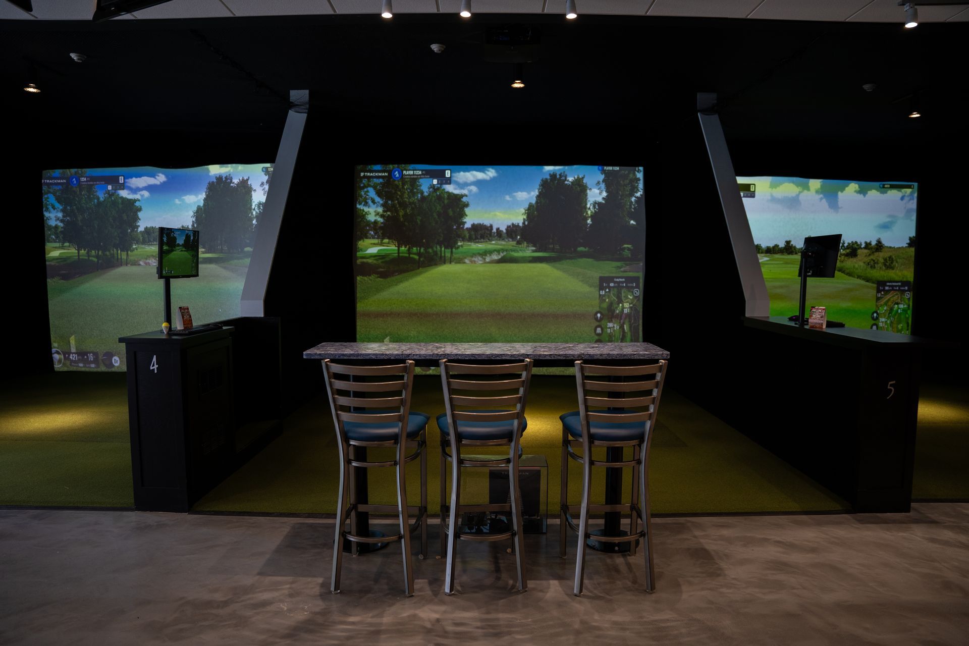 Three indoor golf simulator bays featuring large digital screens, green turf, and high-top chairs with tables.