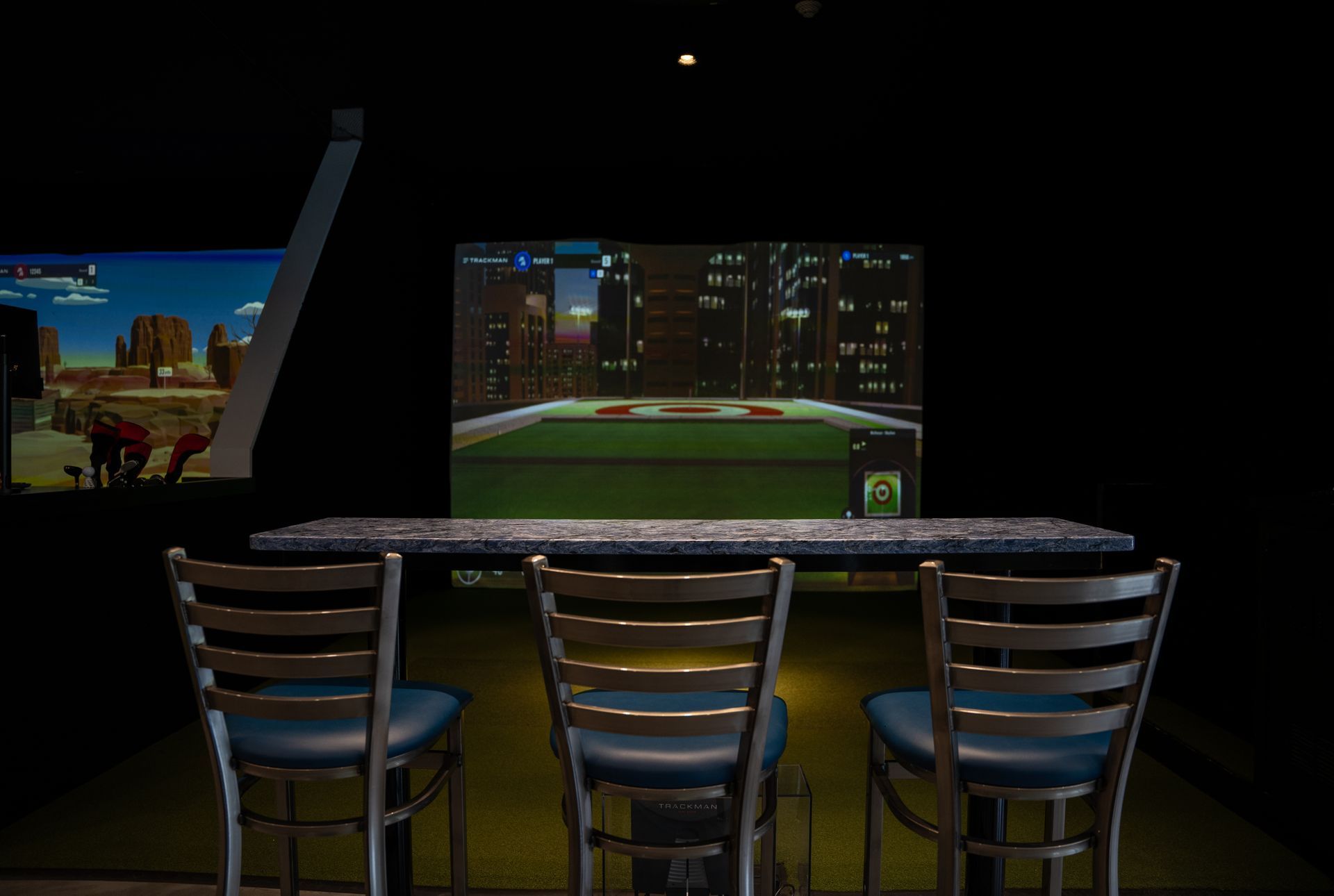 Three chairs face a table in front of a digital golf simulator screen showing a cityscape and a green golf course.