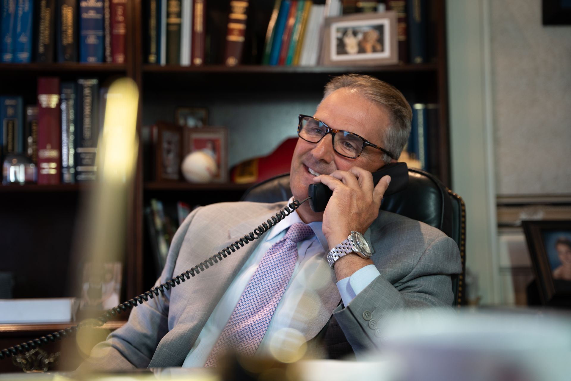 A professional in a suit and glasses sits at a desk in a book-lined office, smiling while talking on a telephone.