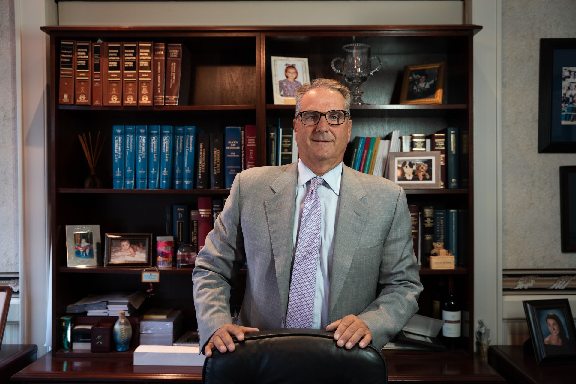 A person in a suit and patterned tie stands behind a desk in an office with a bookshelf filled with books and photos.