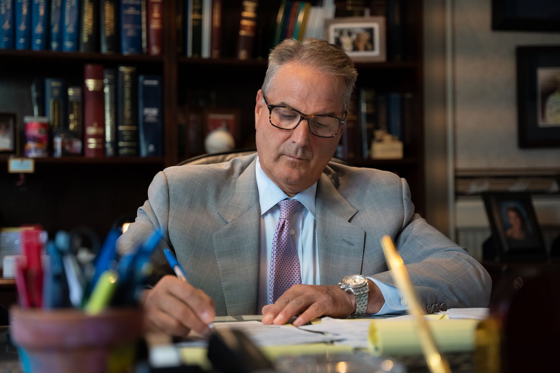 A person in a suit and glasses writing at a desk in an office with bookshelves in the background.