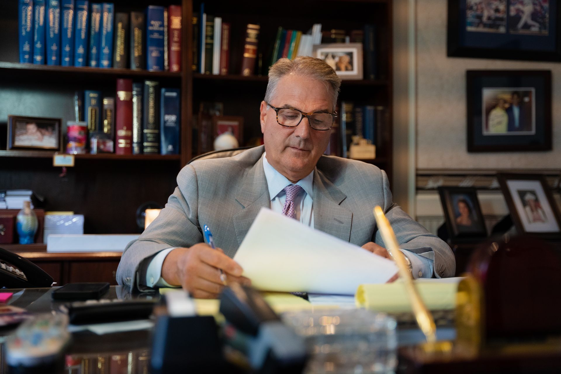 A person in a suit and glasses sits at a desk in a book-filled office, reading a document.