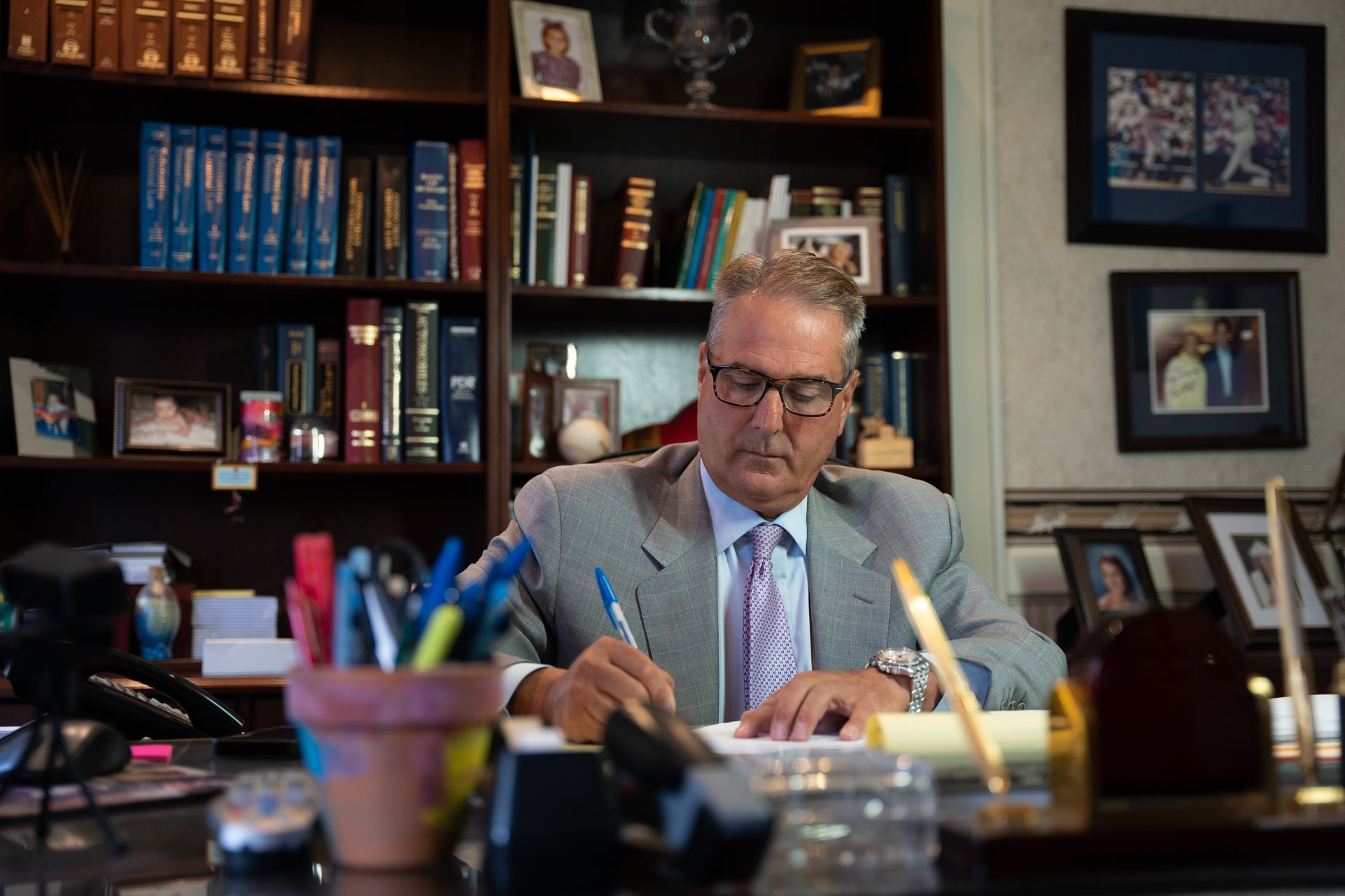 A man in a suit and patterned tie sits at a desk in a book-lined office, writing on a notepad.
